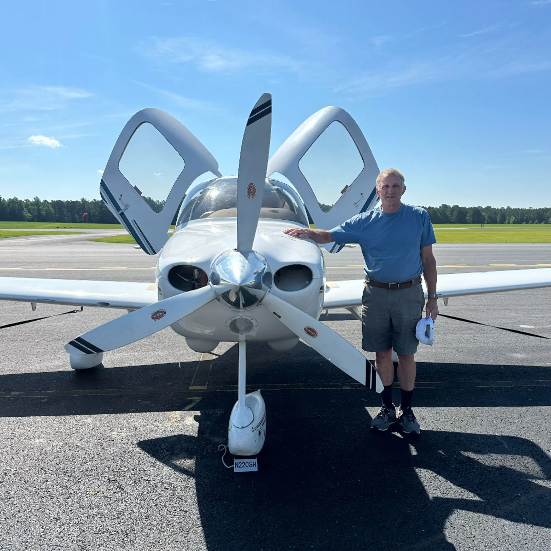 An elderly man in a blue shirt and gray shorts standing next to a white single-engine airplane on a runway, with its doors open and a clear blue sky in the background.