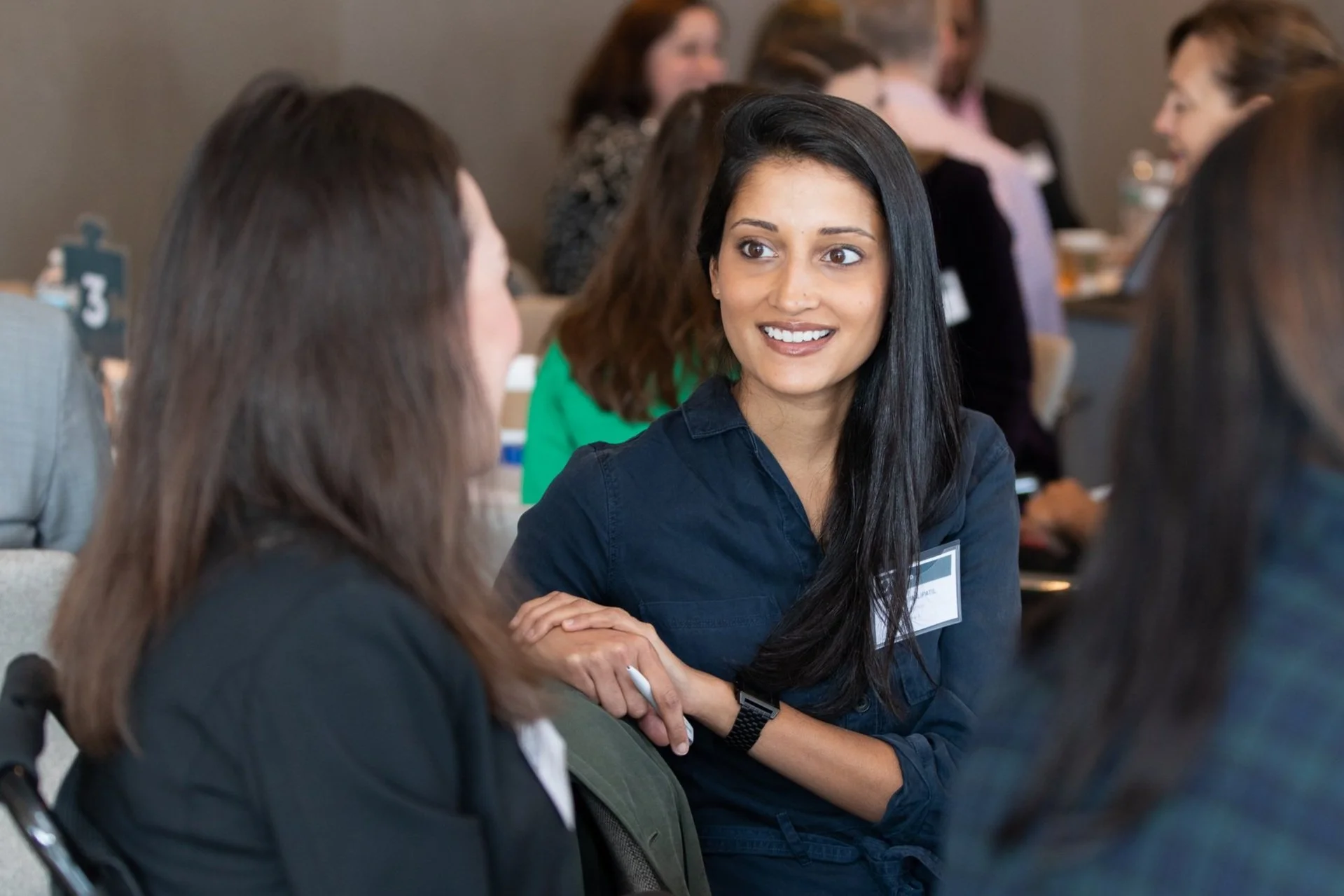 A woman with brown skin and dark hair smiles and looks at another person with long hair, pictured from the back