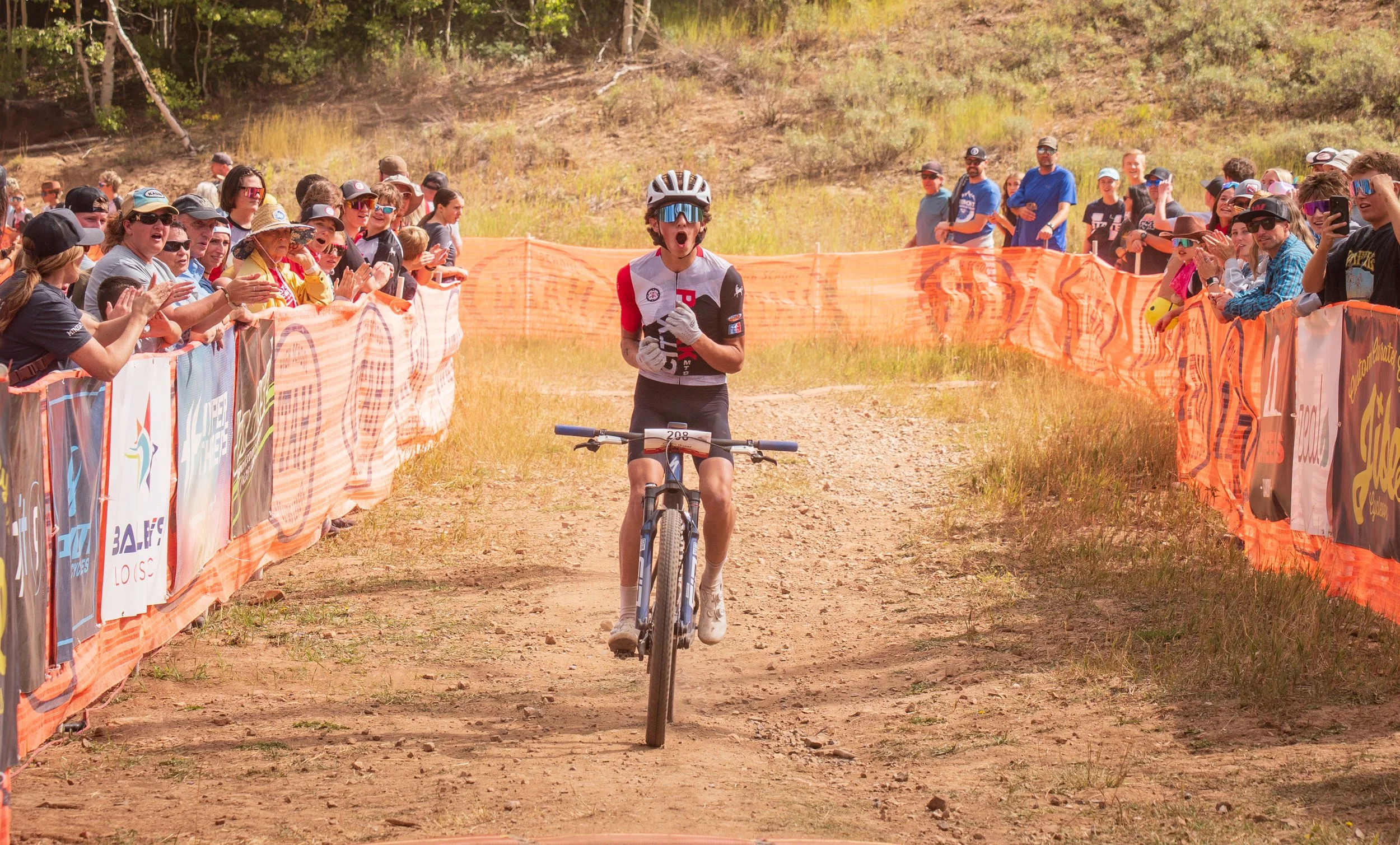 A mountain biker with a helmet and sunglasses crossing a dirt trail during a race, with an excited crowd of spectators clapping and cheering along the sides of the trail.