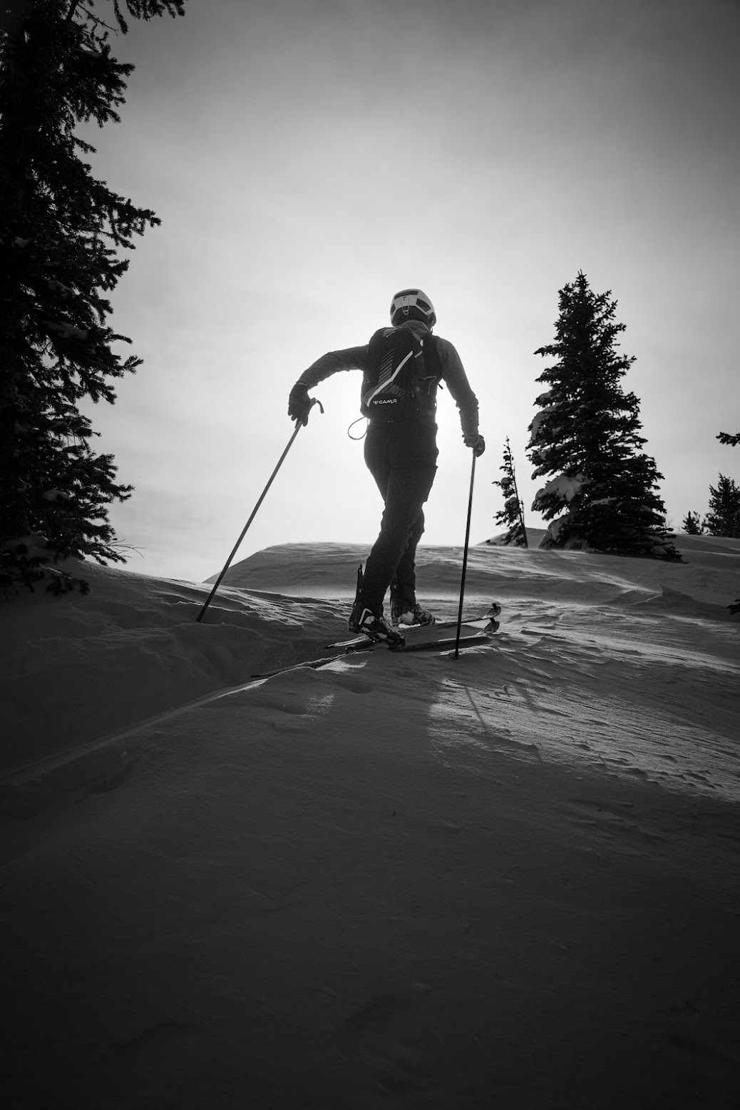 A person skiing on a snowy mountain slope, wearing a helmet, backpack, and skis, surrounded by tall evergreen trees, with the sun shining behind them, creating a silhouette effect in black and white.