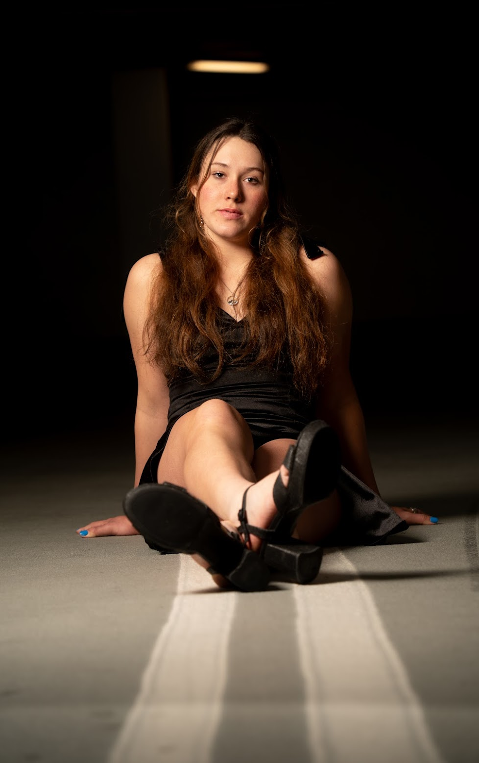A young woman with long brown hair, wearing a black dress and black sandals, sitting on the floor with one leg extended and the other bent, in a dark room under a dim light.