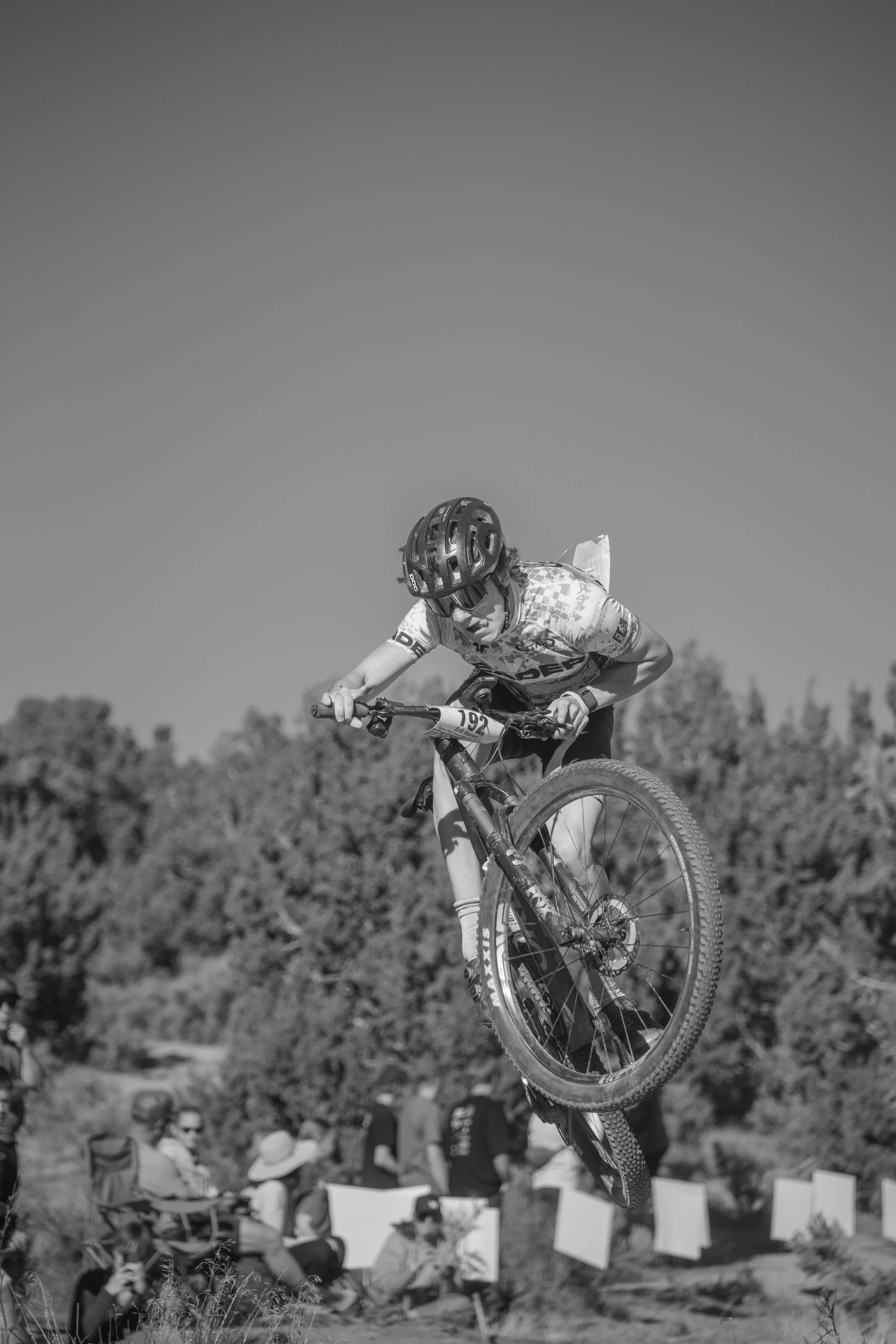 A mountain biker wearing a helmet and sports gear is performing a jump on a dirt trail with a crowd of spectators watching below under a clear sky.