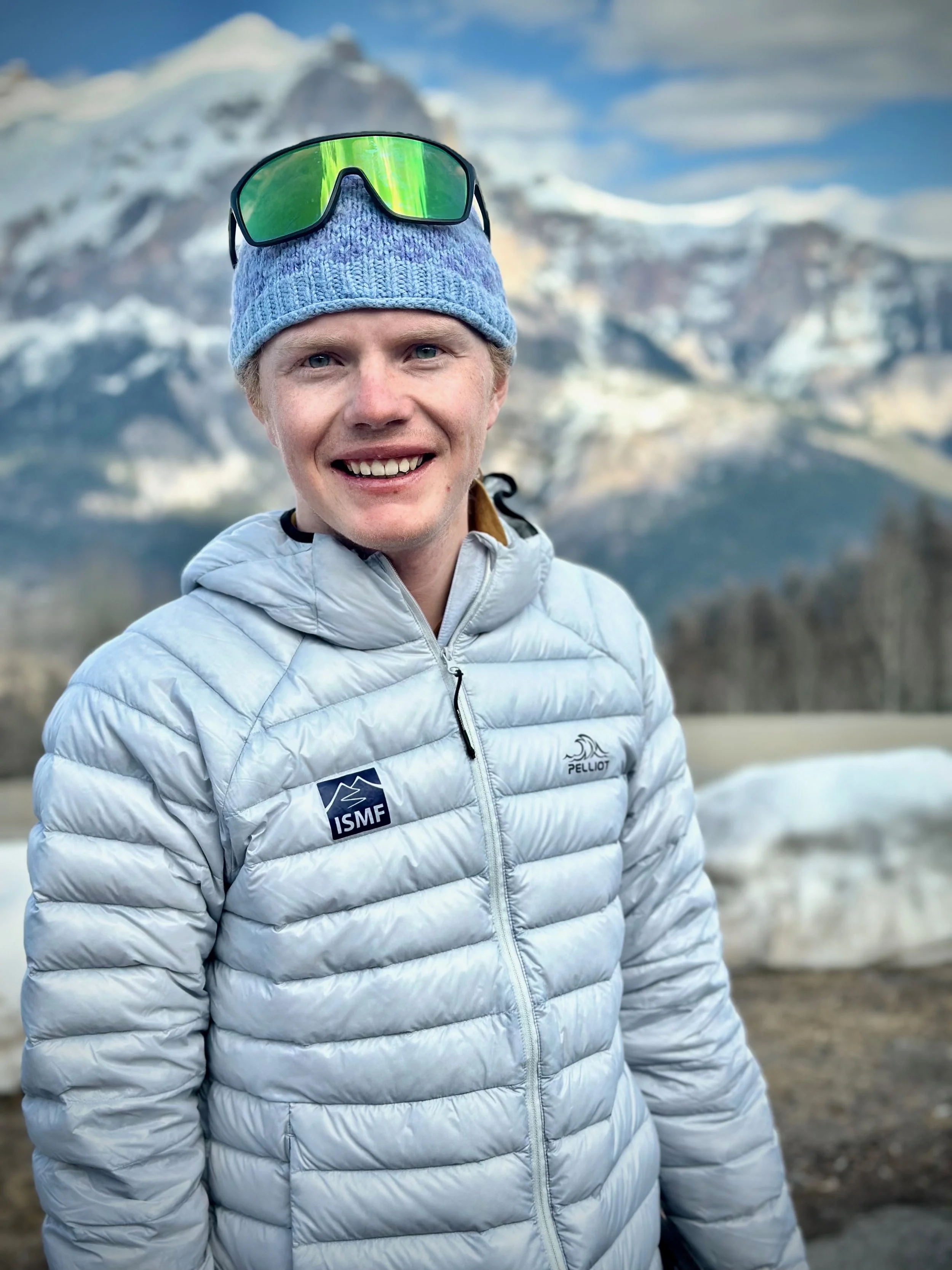 A smiling man wearing a gray winter jacket and a blue knit hat with sunglasses on top, standing outdoors with snow-covered mountains in the background.