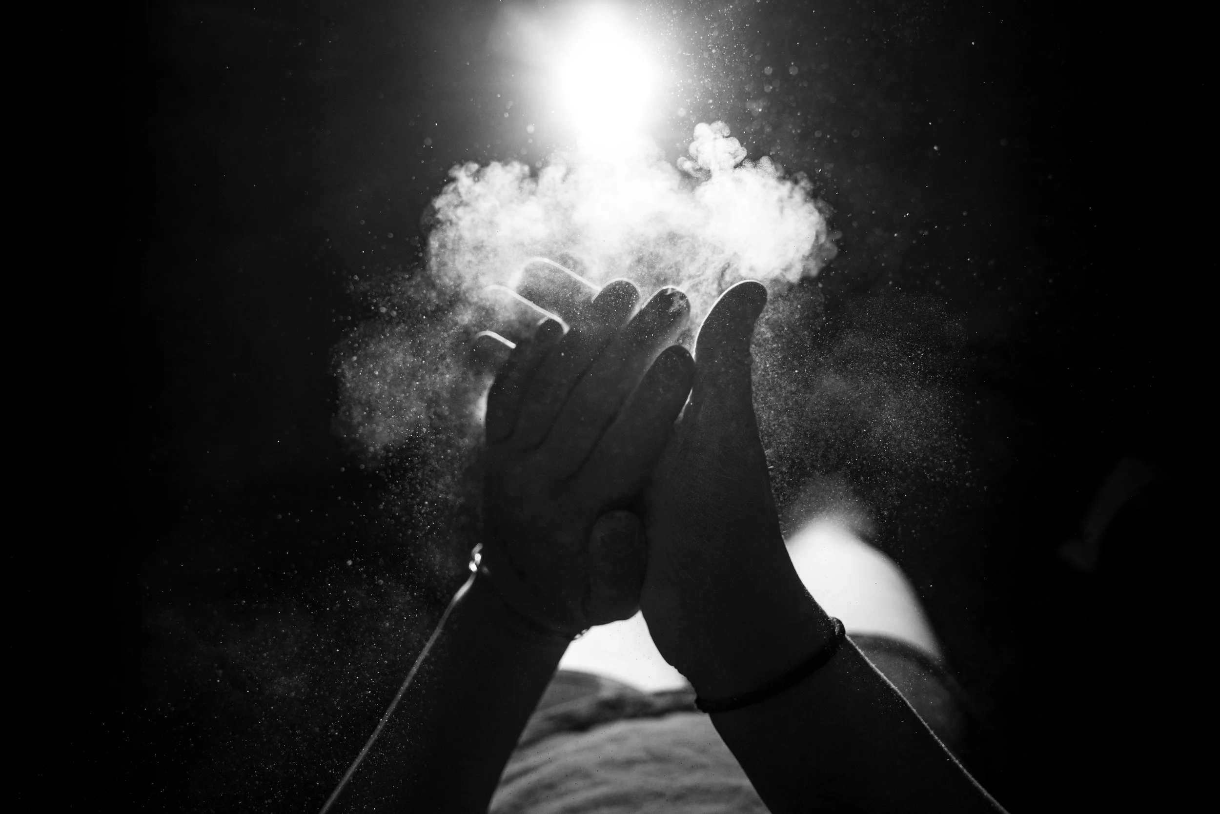 A person's hand is reaching upward, surrounded by dust or powder particles, with bright light shining behind the hand creating a dramatic effect.