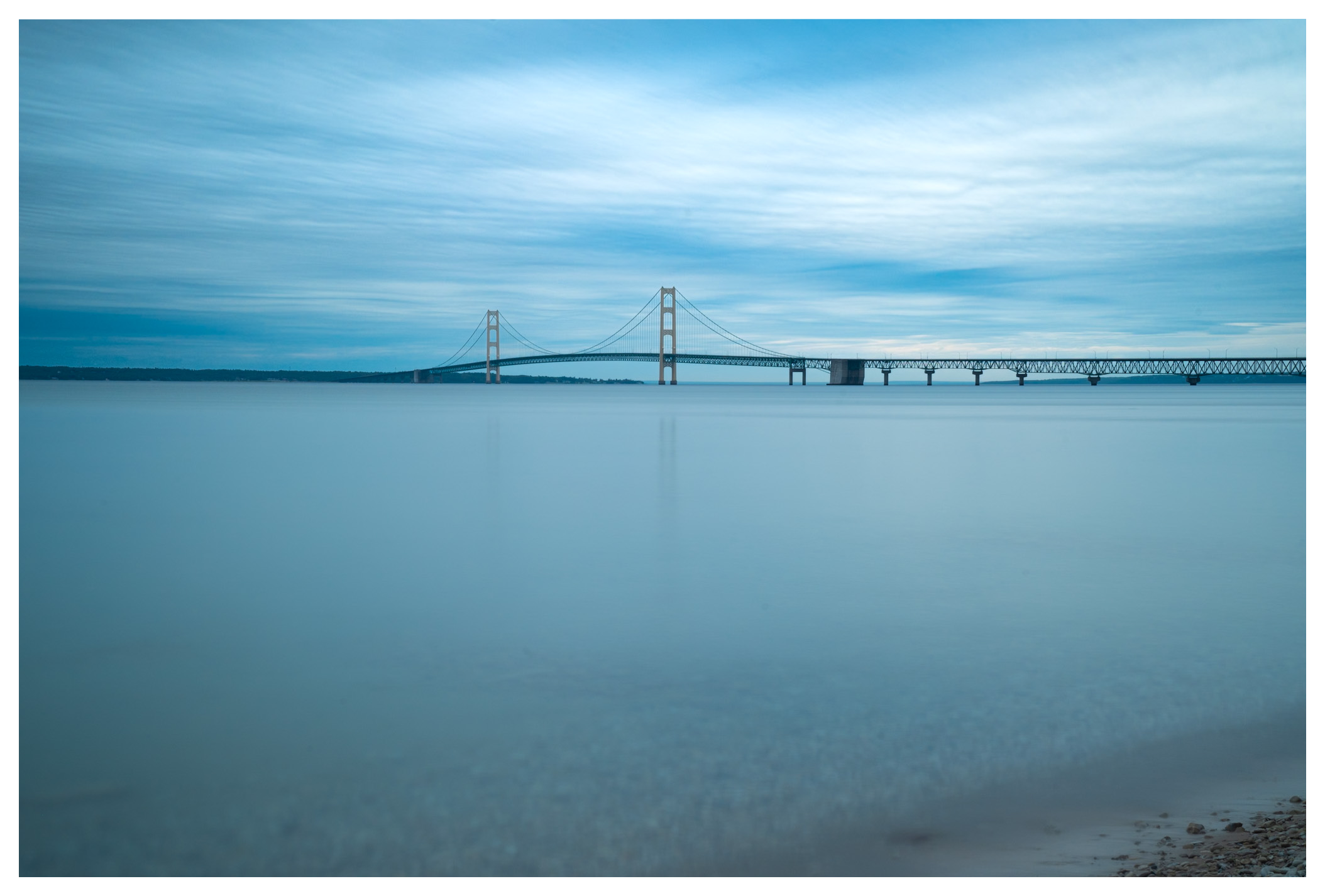 A long suspension bridge over a calm body of water, under a partially cloudy sky.
