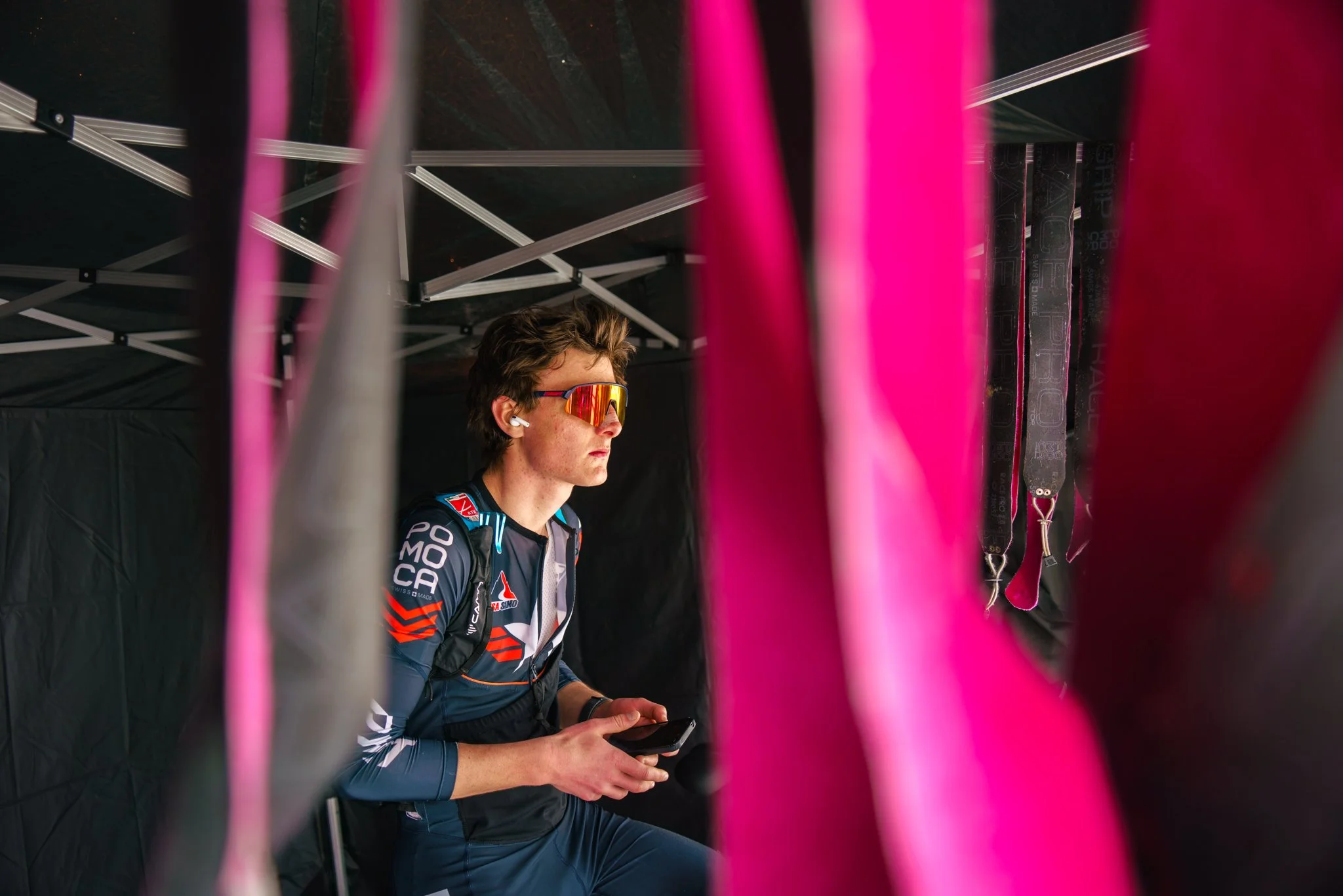 A young male athlete wearing sporty sunglasses, earbuds, and a racing uniform, seated and looking at a device, with colorful flags and tent structure surrounding him.
