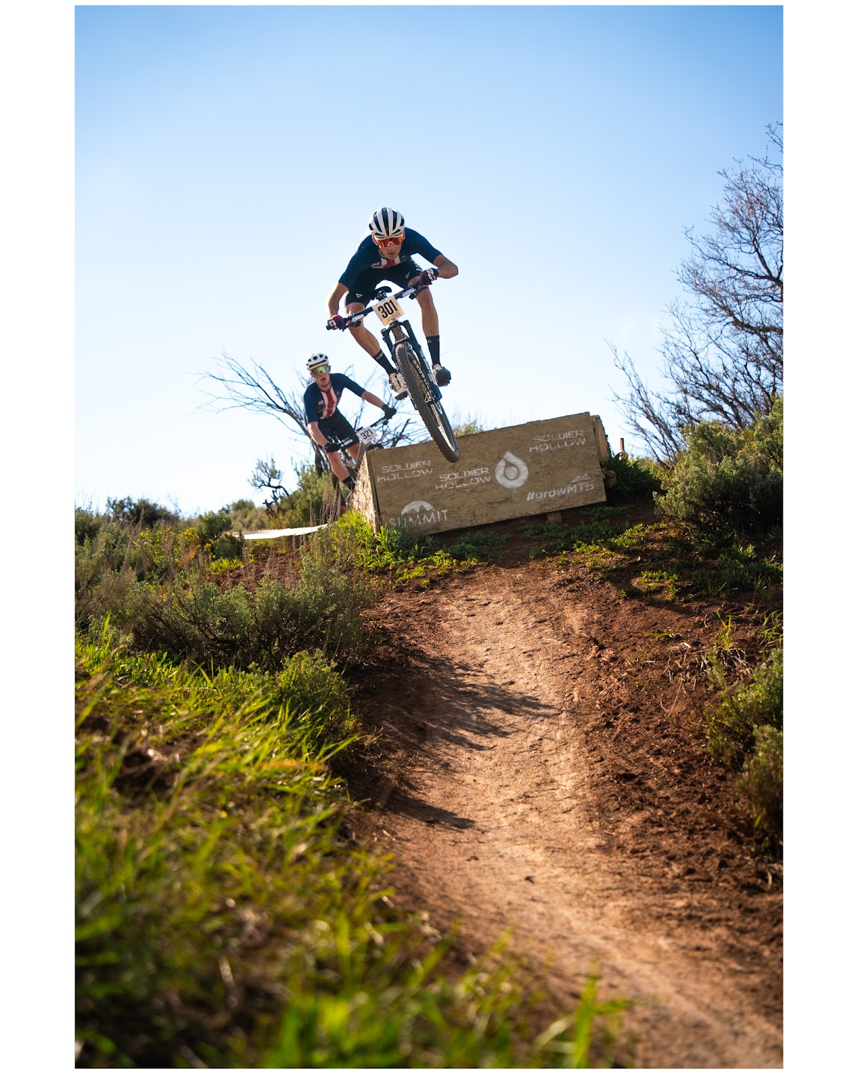 Two mountain bikers wearing helmets and riding gear jump off a dirt trail ledge during a race, with a sign that reads "Soldier Hollow" and hashtag #growwith in the background, under a clear blue sky.