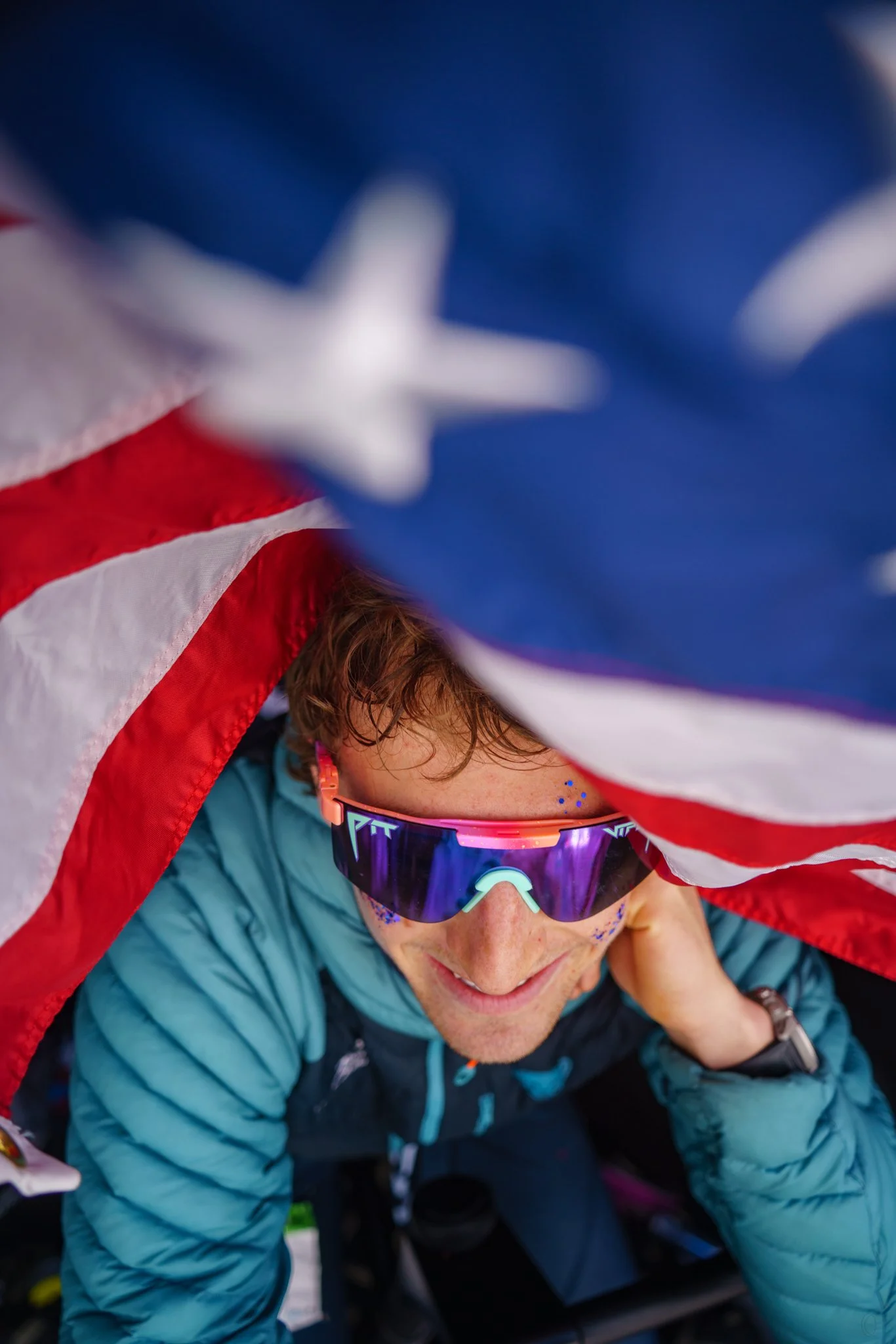 Close-up of a woman with curly hair wearing ski goggles and a blue jacket, holding an American flag above her head.