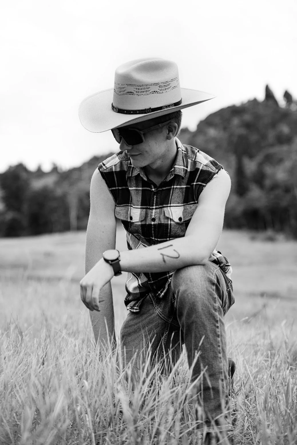 A young man kneeling in a grassy field wearing a sleeveless plaid shirt, sunglasses, a watch, a wide-brimmed hat, and with the number 12 marked on his left arm. The photo is in black and white.