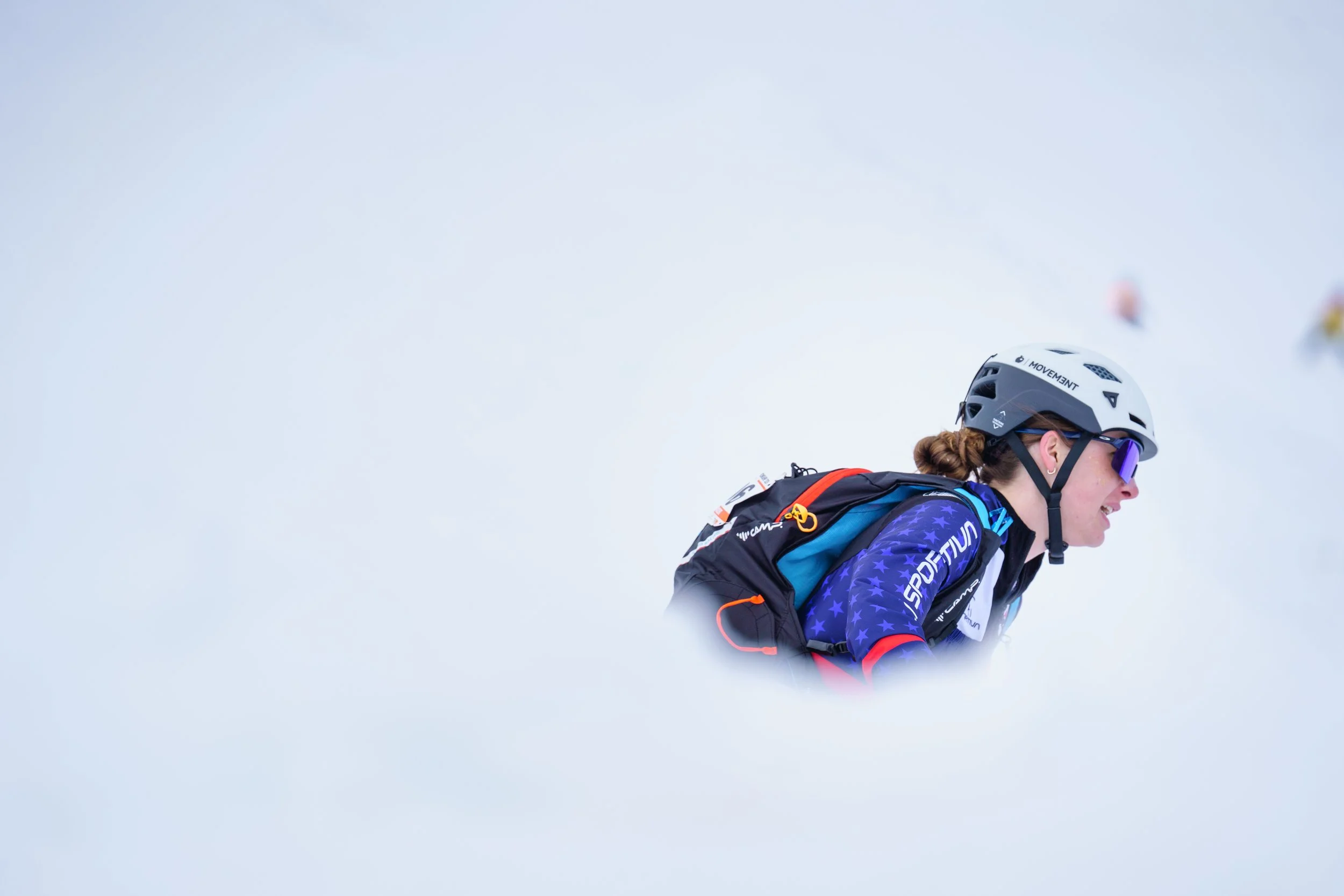 A woman wearing skiing gear, including a helmet, goggles, and a jacket, is smiling while skiing in a snowy environment.