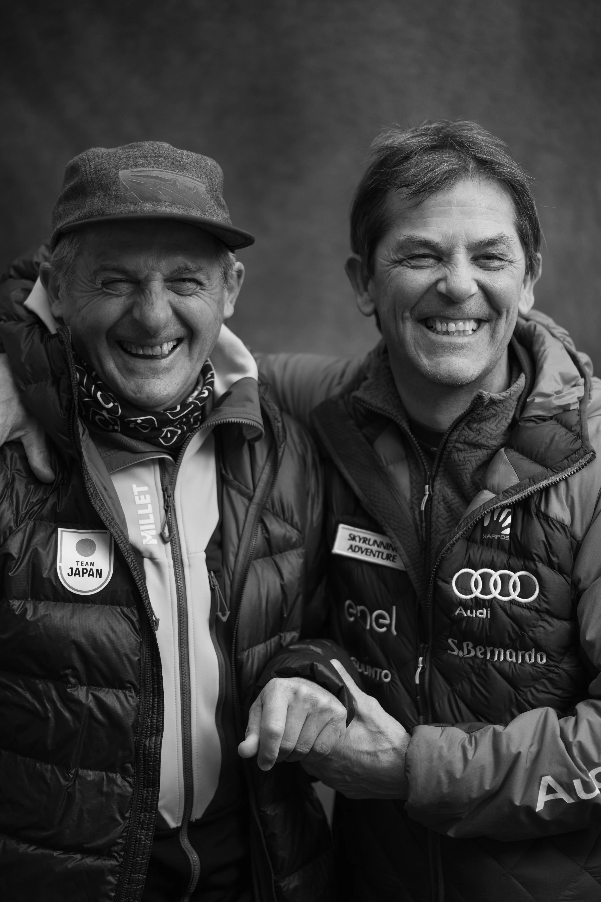 Two smiling men wearing outdoor jackets, one with a cap, standing close together and holding hands, with badges indicating they are part of Team Japan and involved in skyrunning adventure.