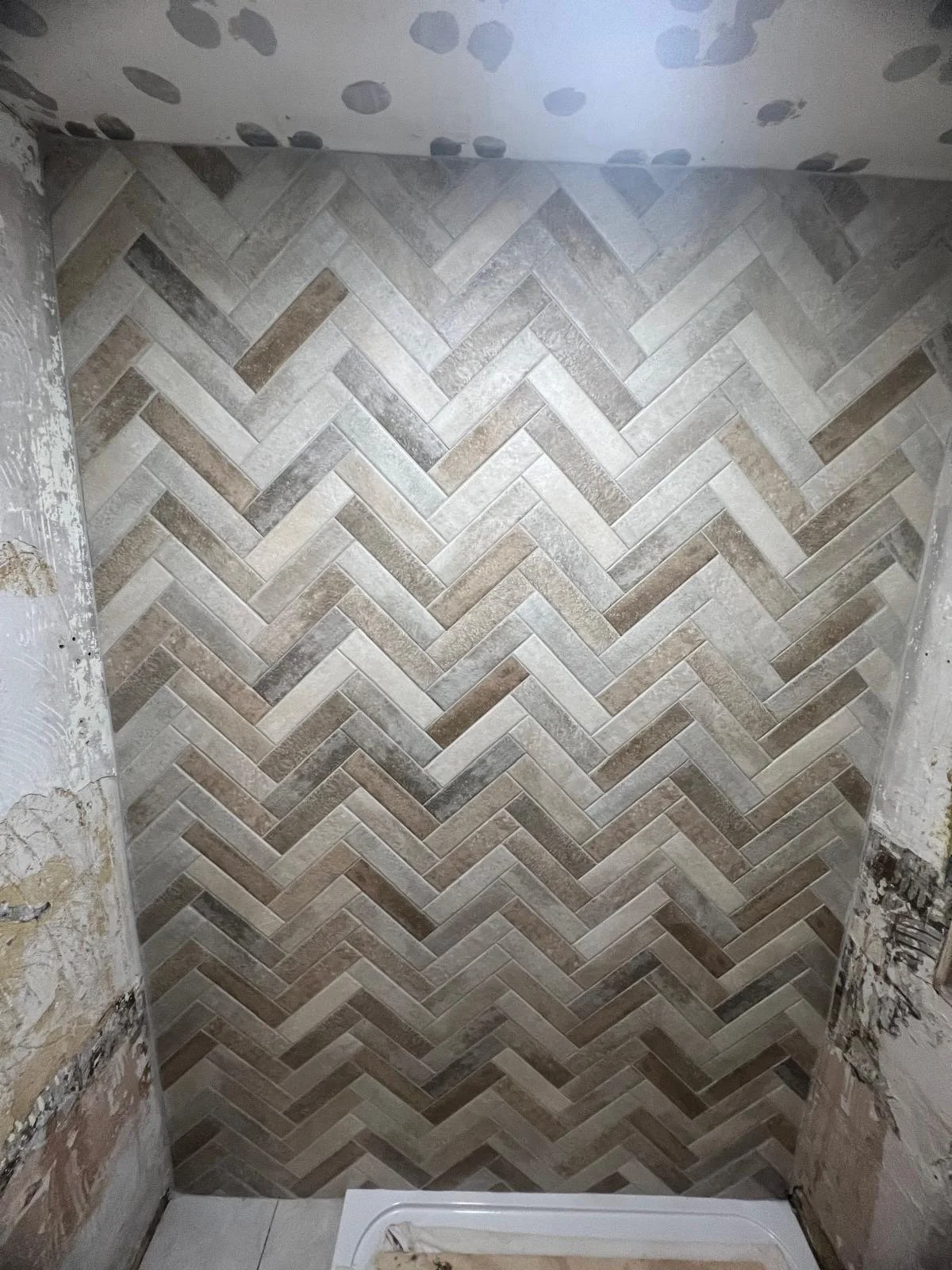Bathroom wall with a herringbone pattern of beige, brown, and gray tiles, some sections of the wall are unfinished with exposed drywall and plaster.