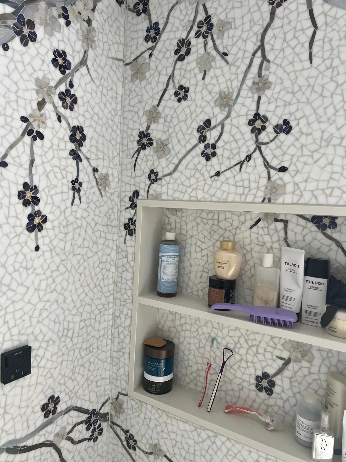 Bathroom shelf with travel-size bottles, skincare products, toothbrushes, and a razor against a white mosaic wall with black floral pattern.