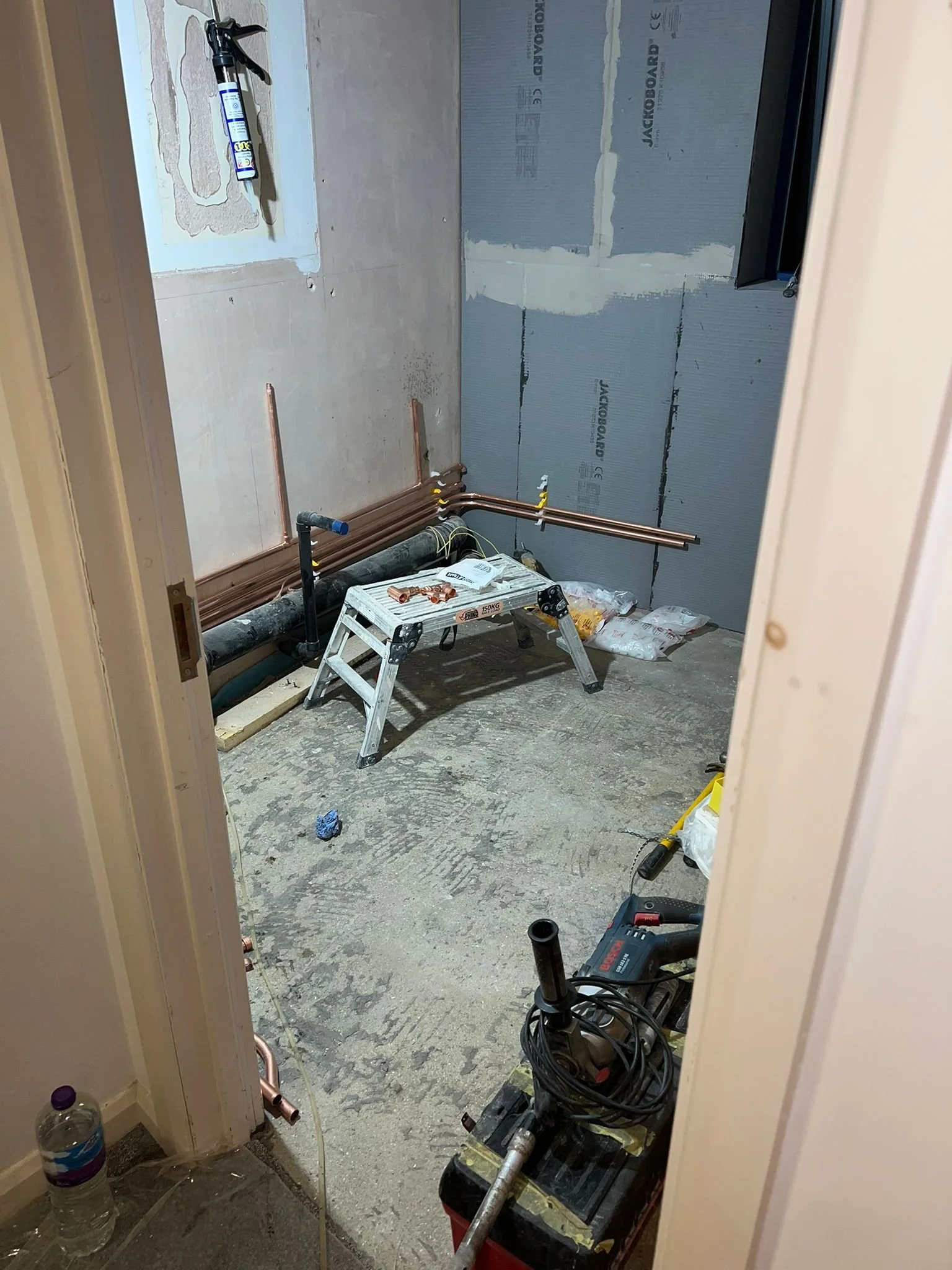 A room under construction with exposed copper pipes, construction tools, a level, a water bottle, and unfinished drywall on the walls.