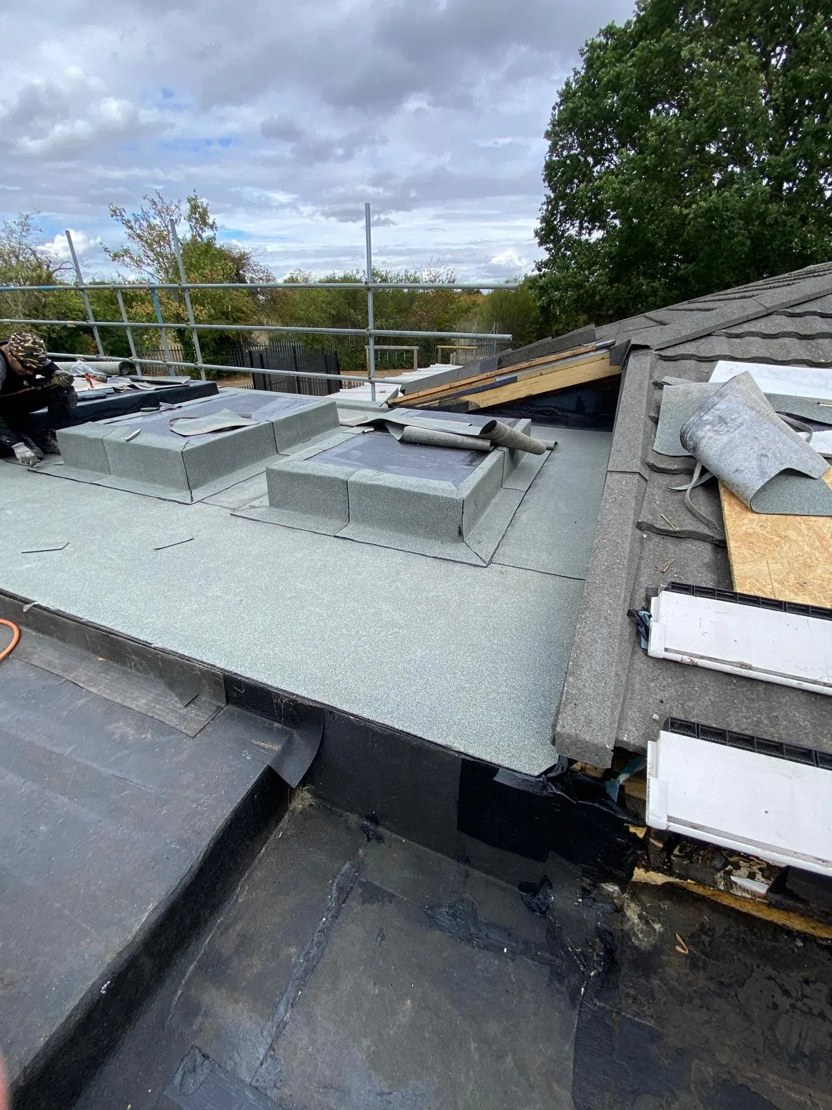 Construction workers installing new roofing on a house with gray shingles, ventilation units, and materials scattered on the roof, surrounded by trees and partly cloudy sky.