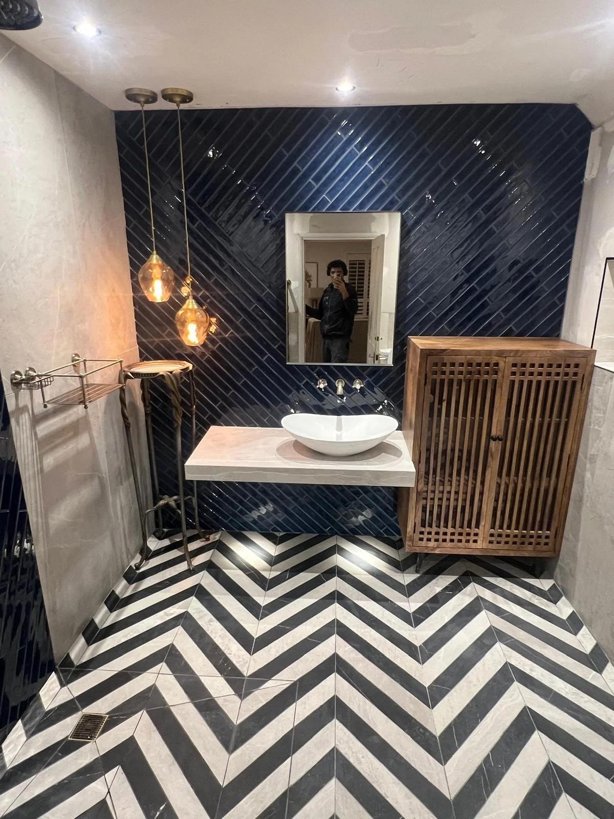 Modern bathroom with black diagonal tiled accent wall, black and white chevron patterned floor, white vessel sink on a light-colored countertop, mirror, wooden cabinet, and pendant lights.