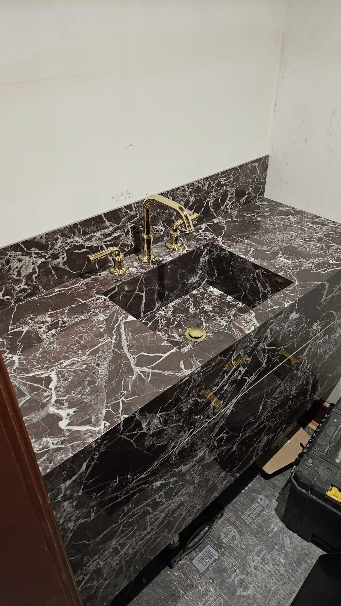 A marble bathroom vanity with a gold faucet, two handles, and a sink, under construction against a white wall.