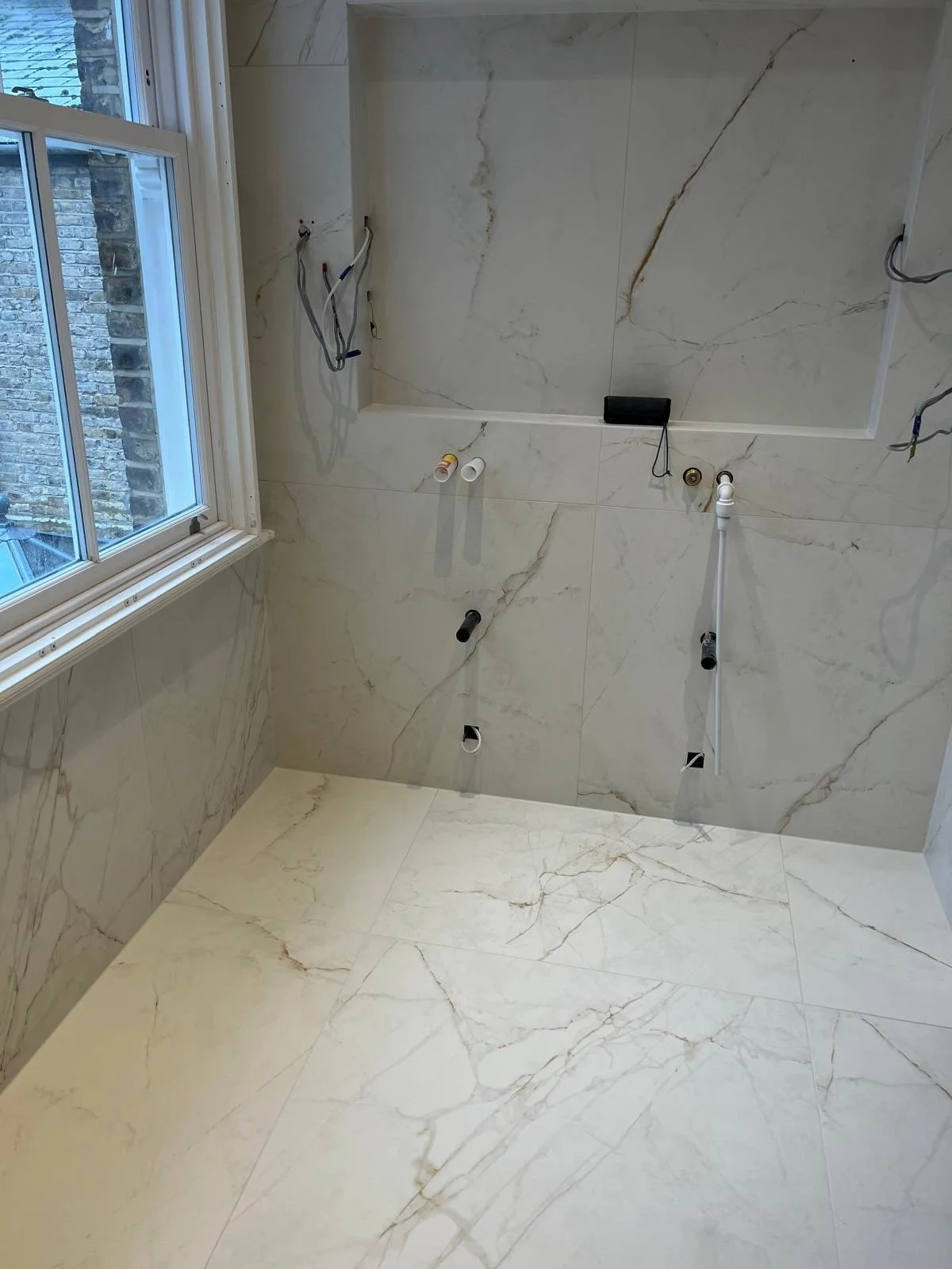 Bathroom with marble tiles on walls and floor, window on the left, exposed plumbing and electrical wiring for a sink or vanity yet to be installed.