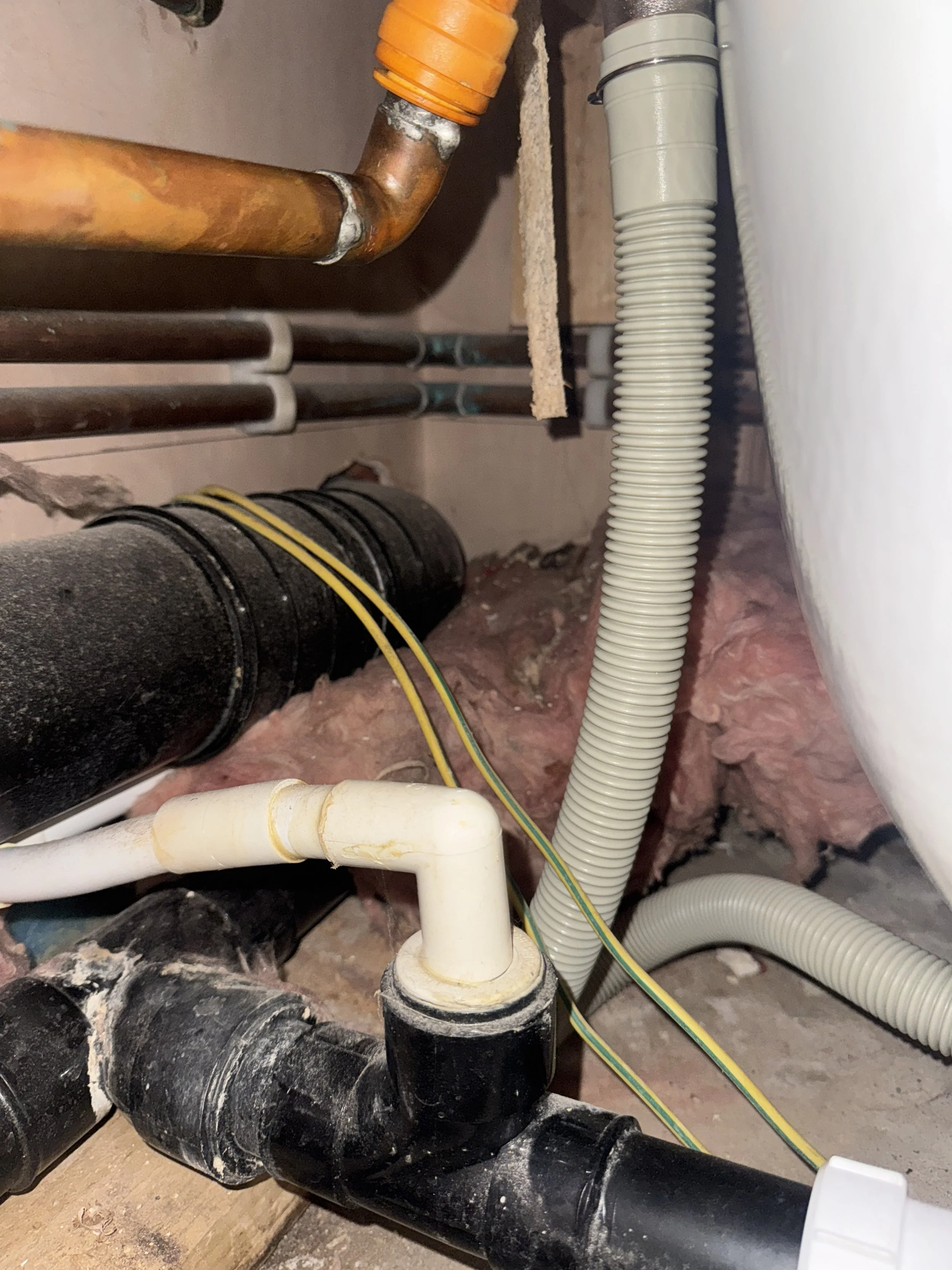 A close-up view of plumbing pipes and tubing under a sink or toilet, including black, white, and metallic pipes and flexible tubing, with some corrosion and dusty debris.