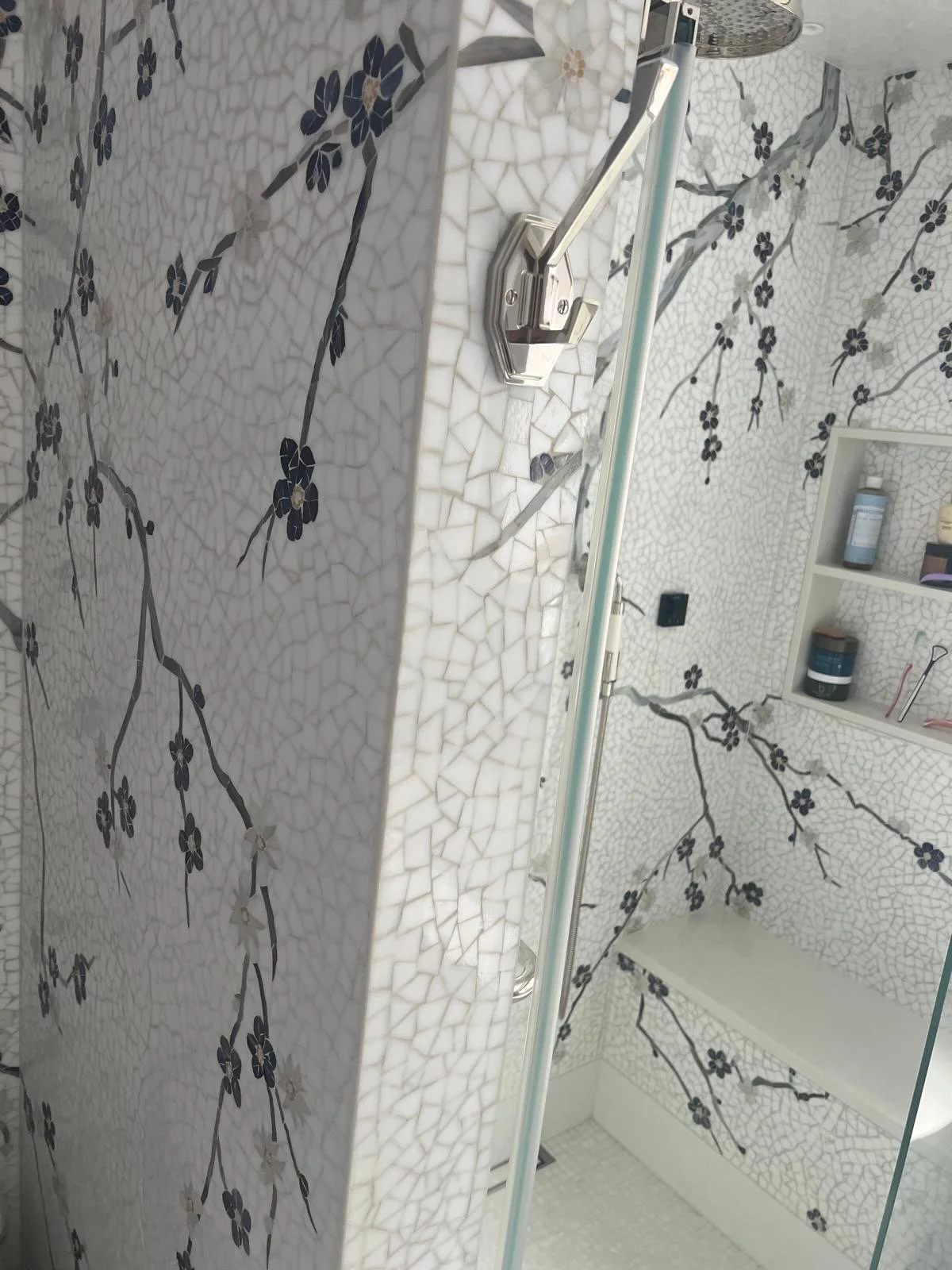 Bathroom shower with white mosaic tiles featuring black floral vine patterns, a glass shower door, and a built-in shelf with toiletries.