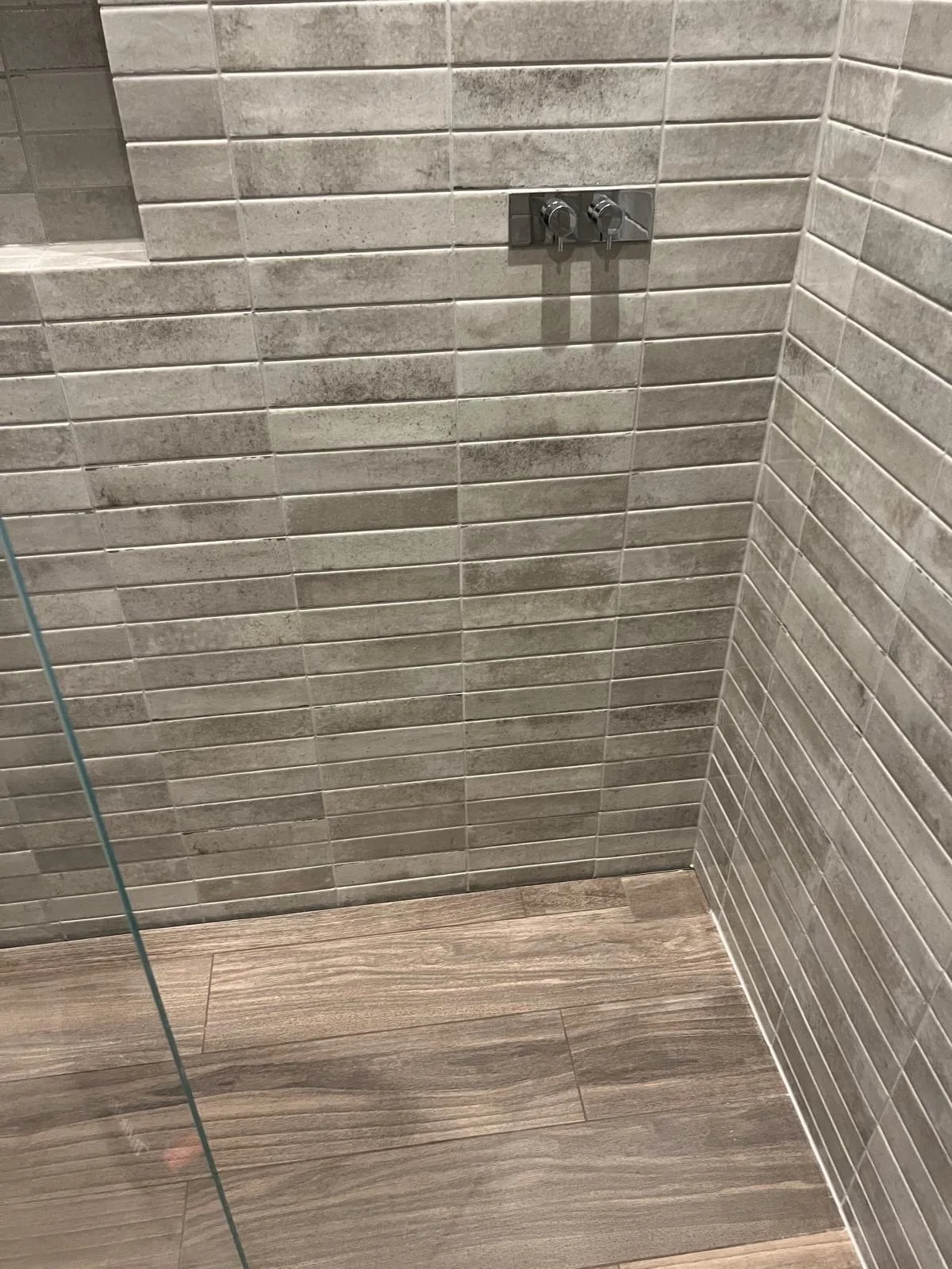 Close-up of a shower wall with gray rectangular tiles, a metallic fixture with two knobs, and wooden-patterned floor tiles.