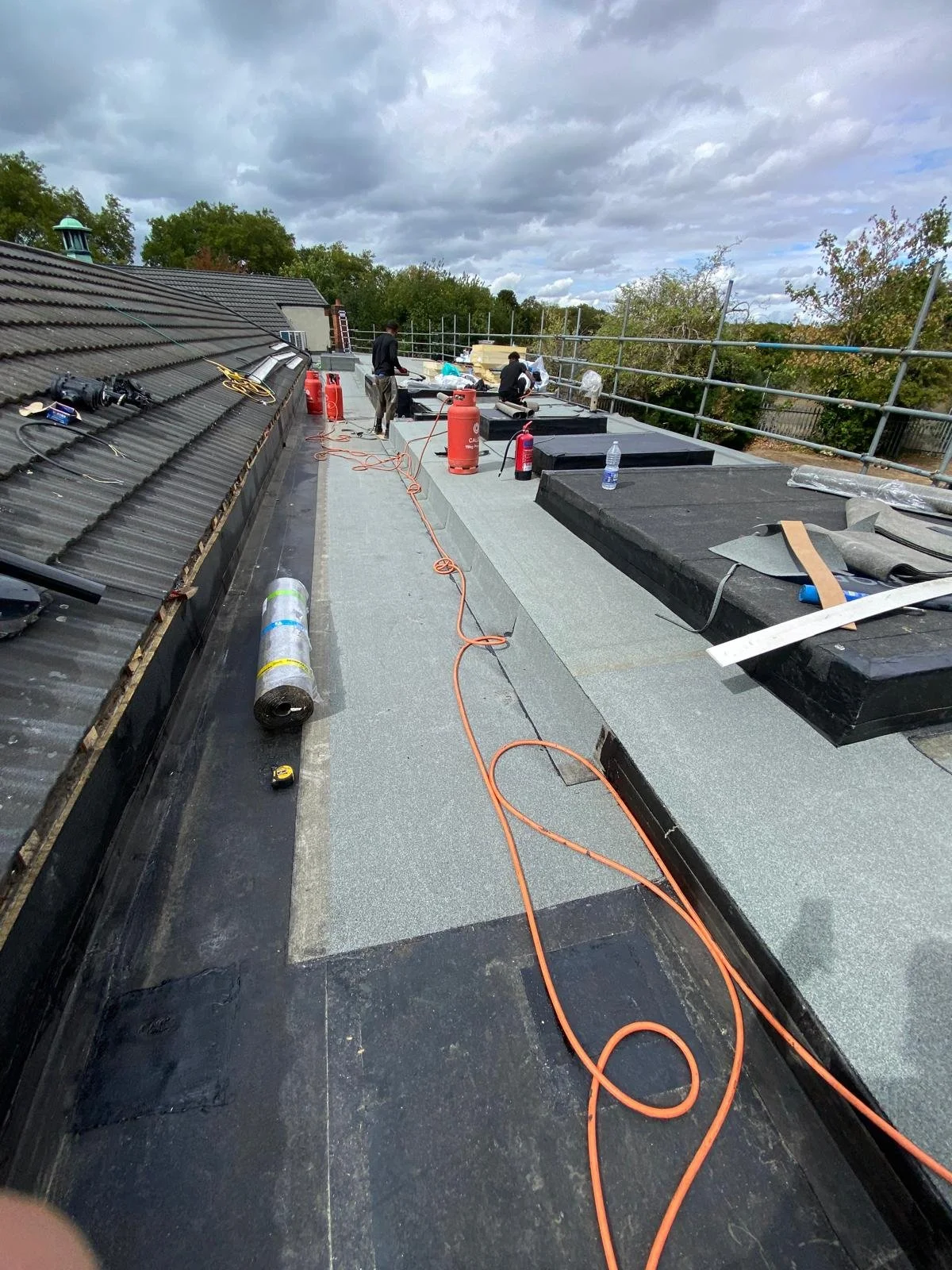 Construction workers on a rooftop working with tools and safety equipment, with cloudy sky overhead.
