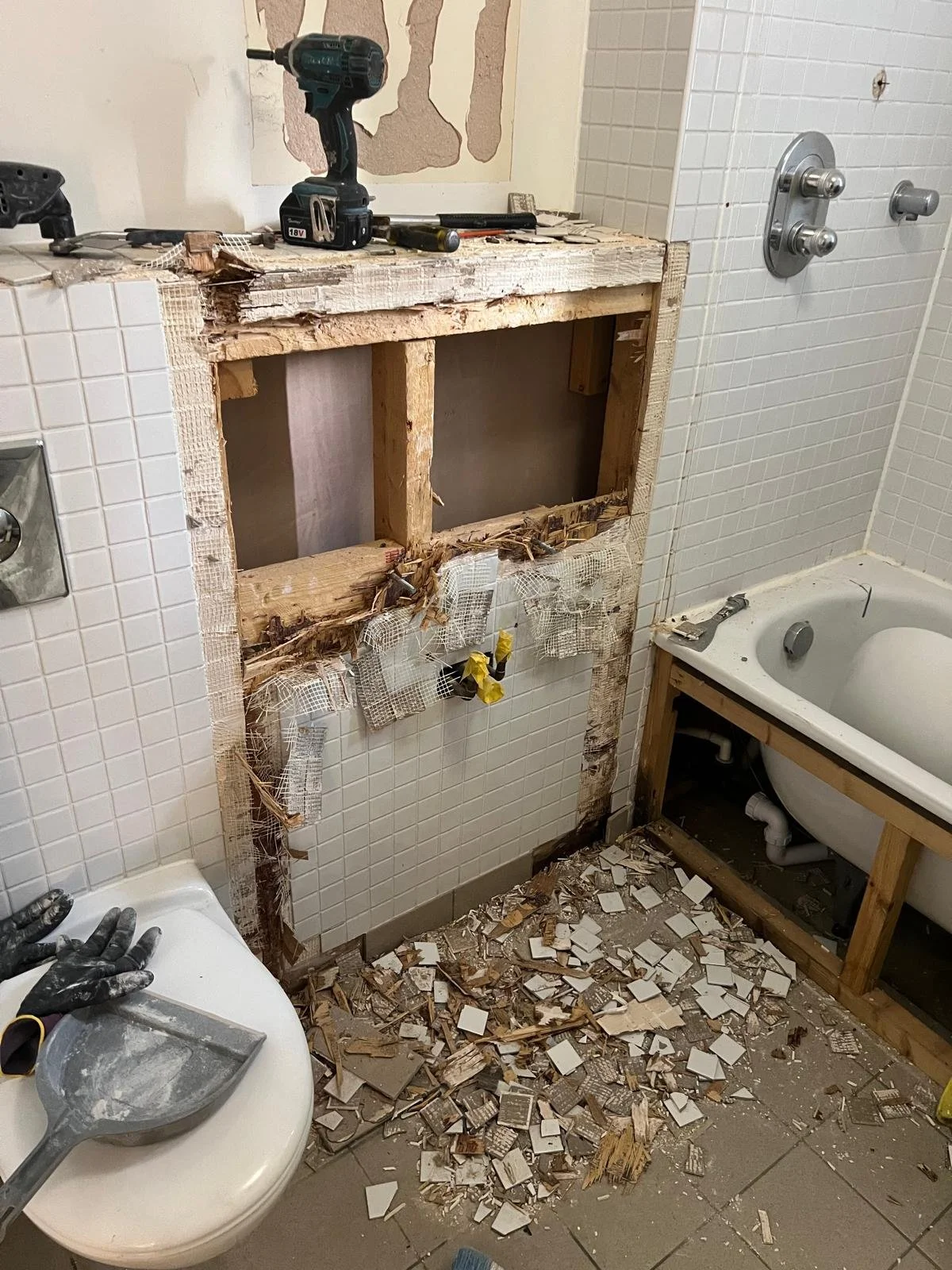 A bathroom under renovation with tiles removed from wall and a large hole in the wall exposing studs, debris and broken tiles scattered on the floor, tools on the sink and a workbench.