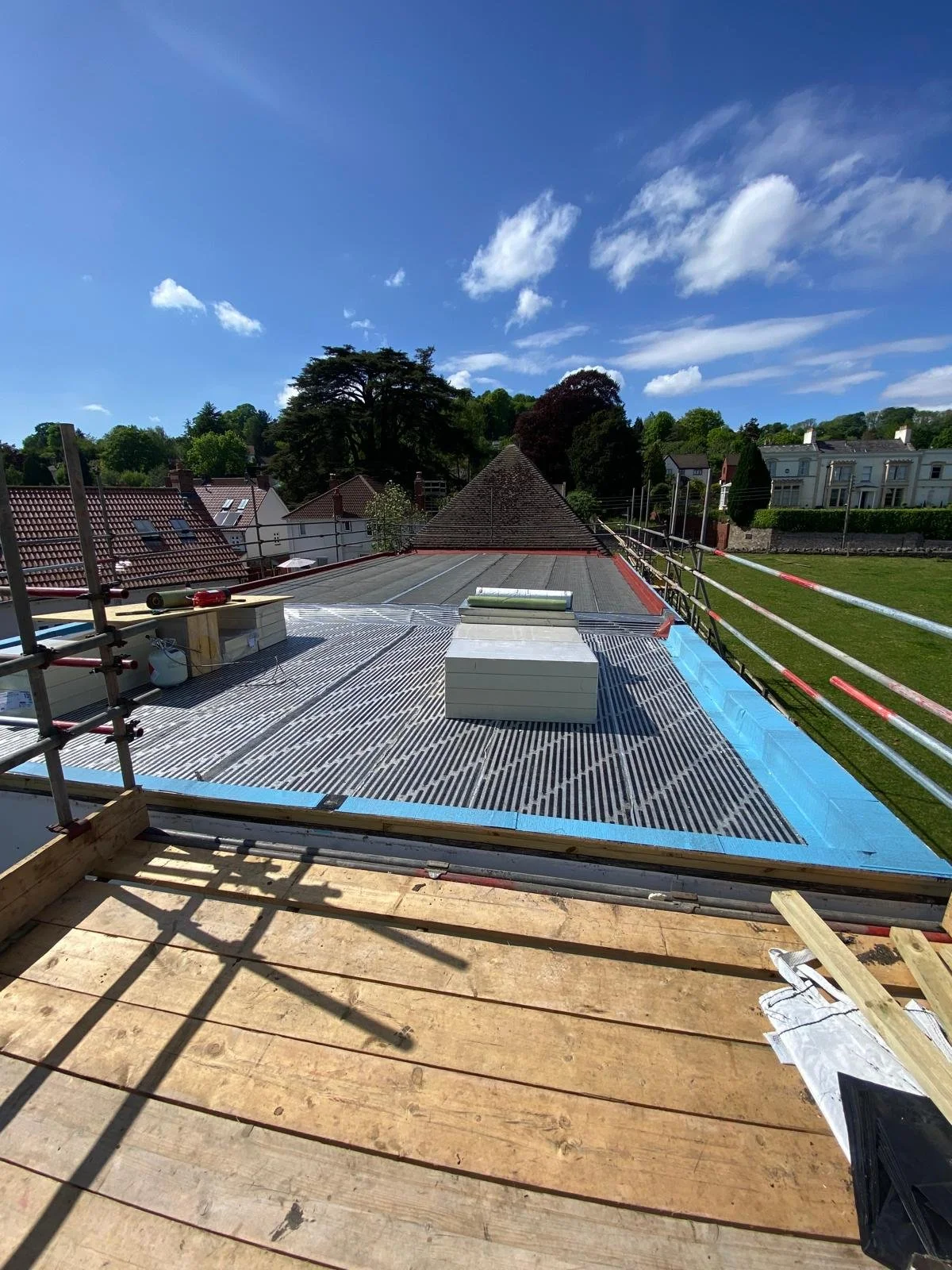View of a rooftop under construction with scaffolding, wooden planks, and roofing materials, surrounded by trees and neighboring houses, under a blue sky with some clouds.