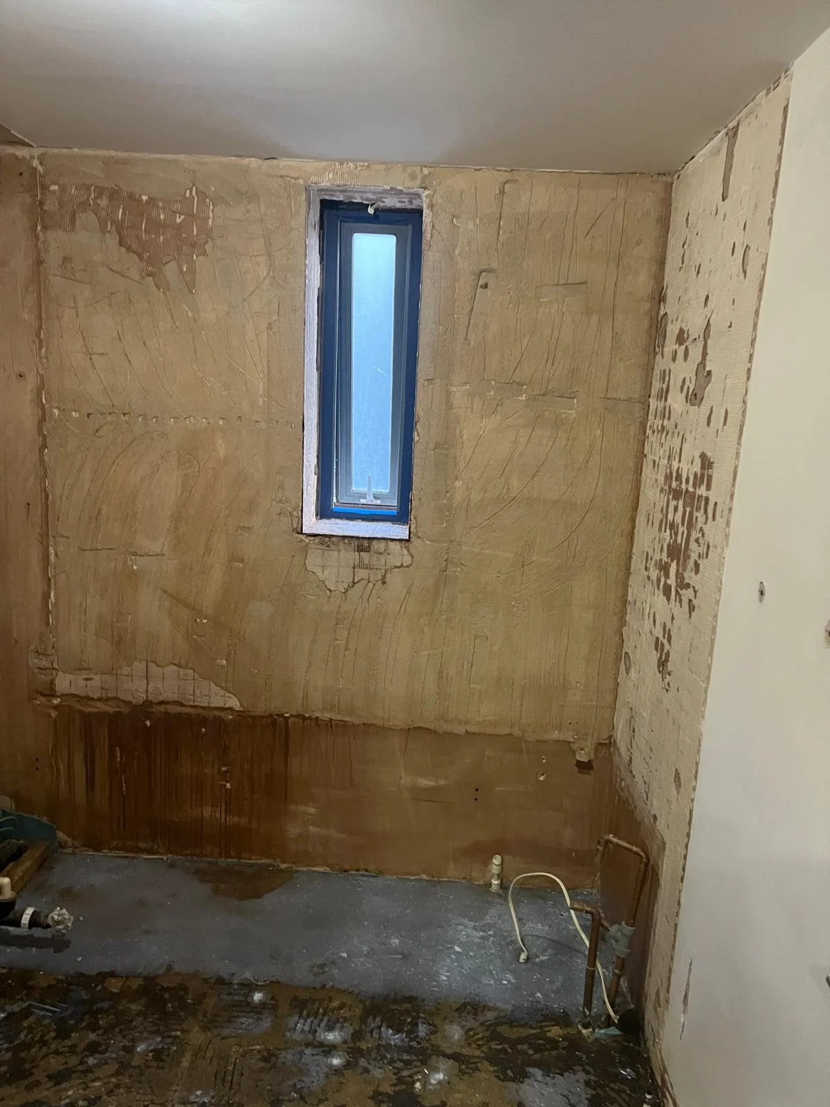A room under renovation with partially removed wall coverings, a narrow window, exposed plumbing, and a stained floor.