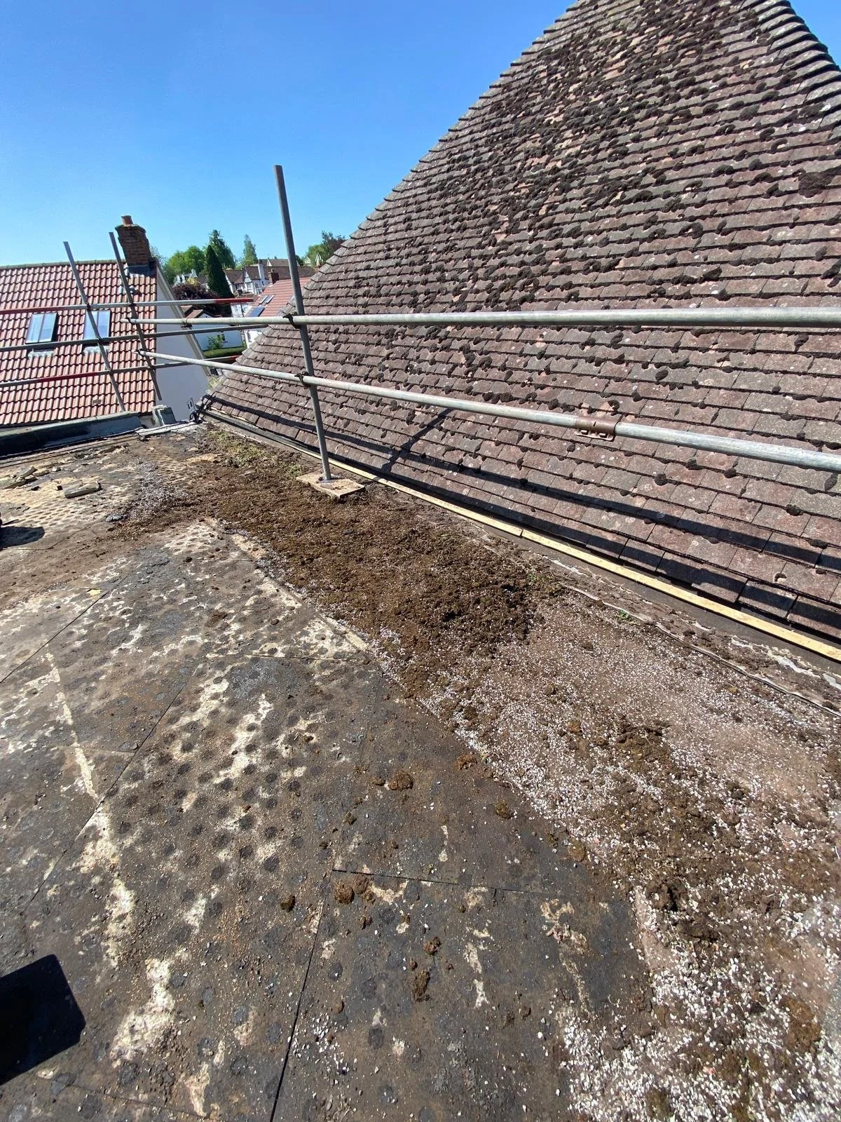 View of a roof with scaffolding and weathered, mossy tiles, adjacent to a flat surface with dirt and debris, under clear blue sky.