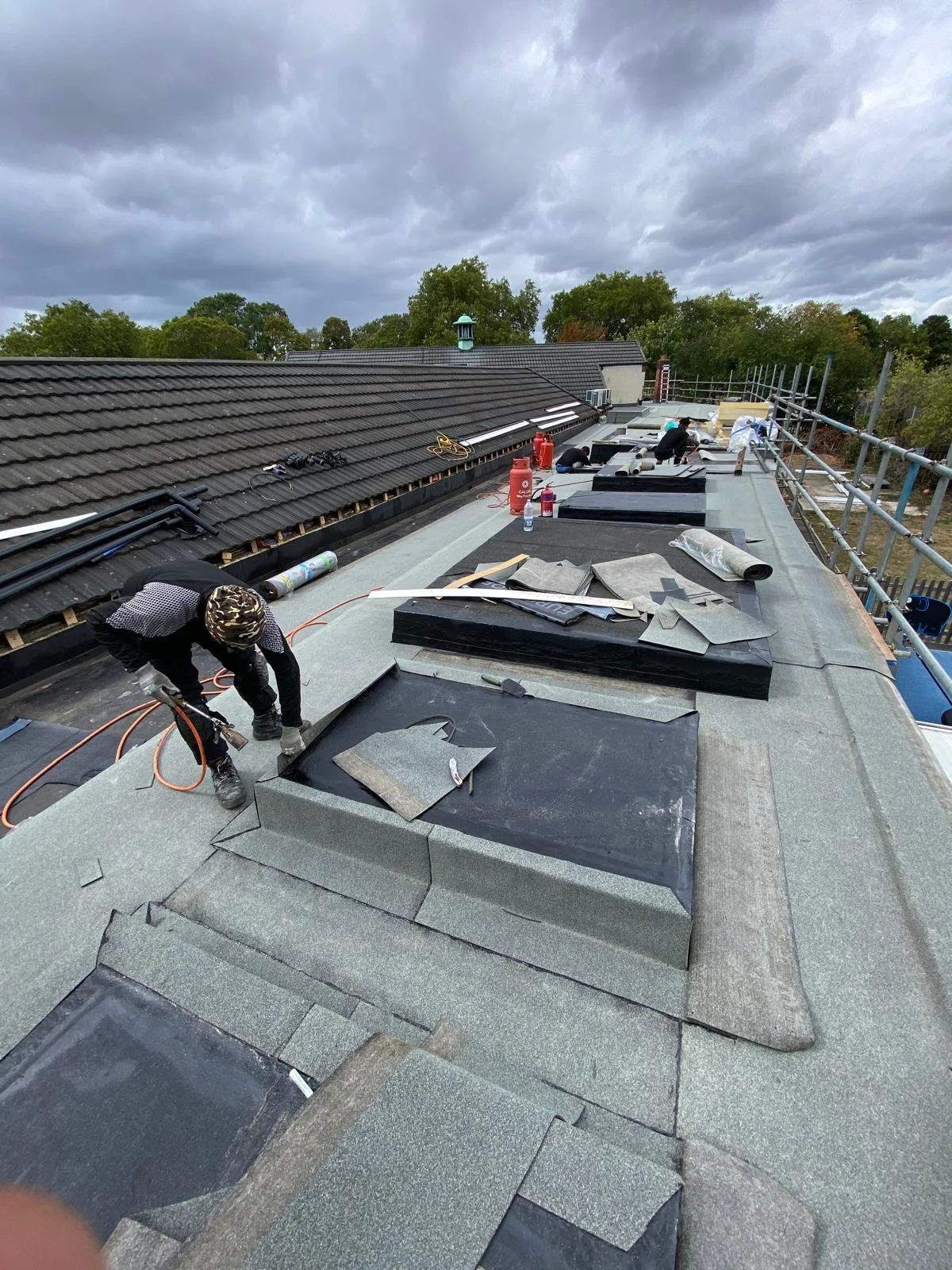 Roofers installing roofing materials on a residential roof under cloudy skies, with trees in the background.