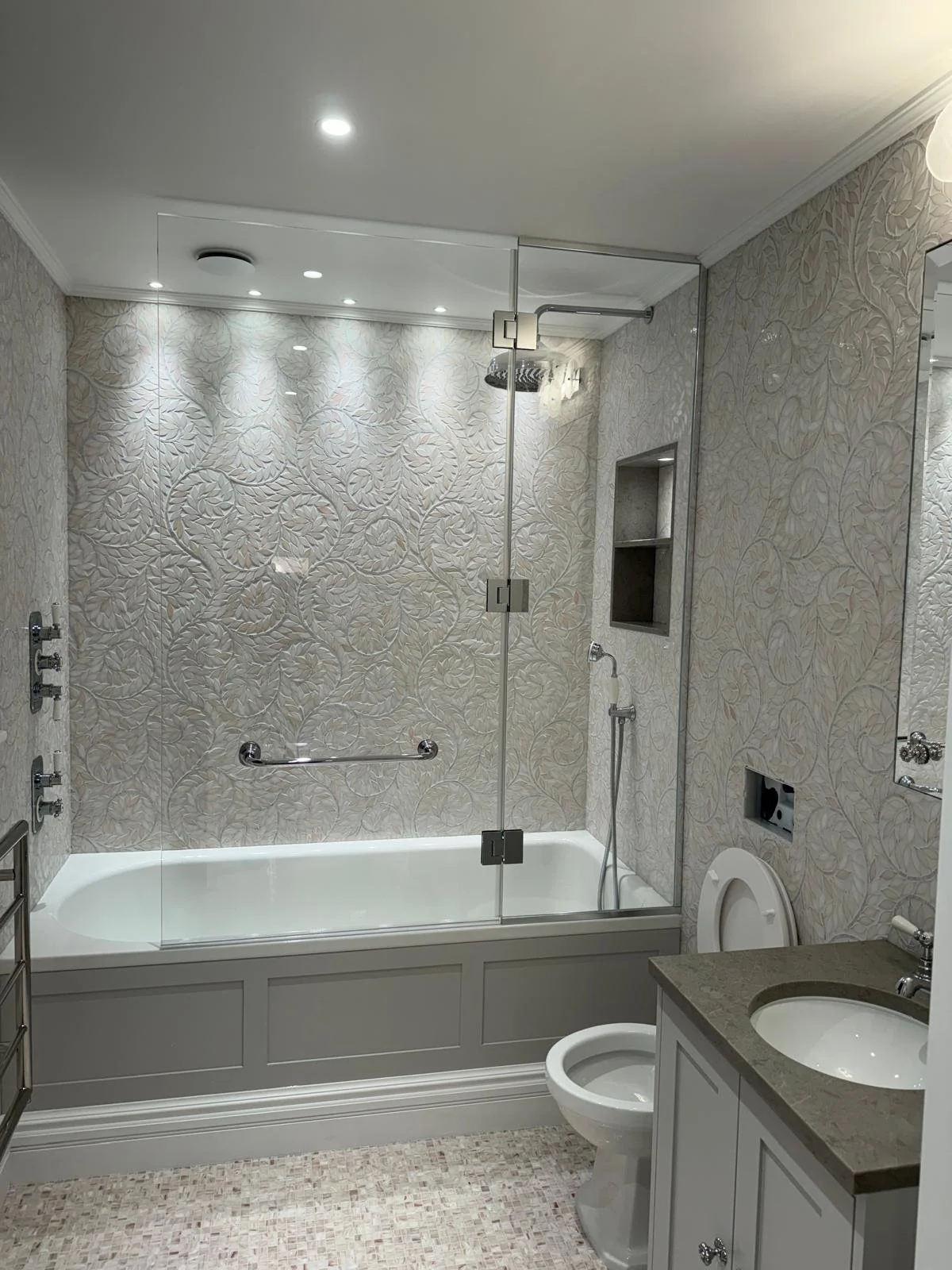 A bathroom with a bathtub that has a glass shower enclosure, a textured floral wallpaper, a small niche in the wall, and a white vanity with a granite countertop and a sink.