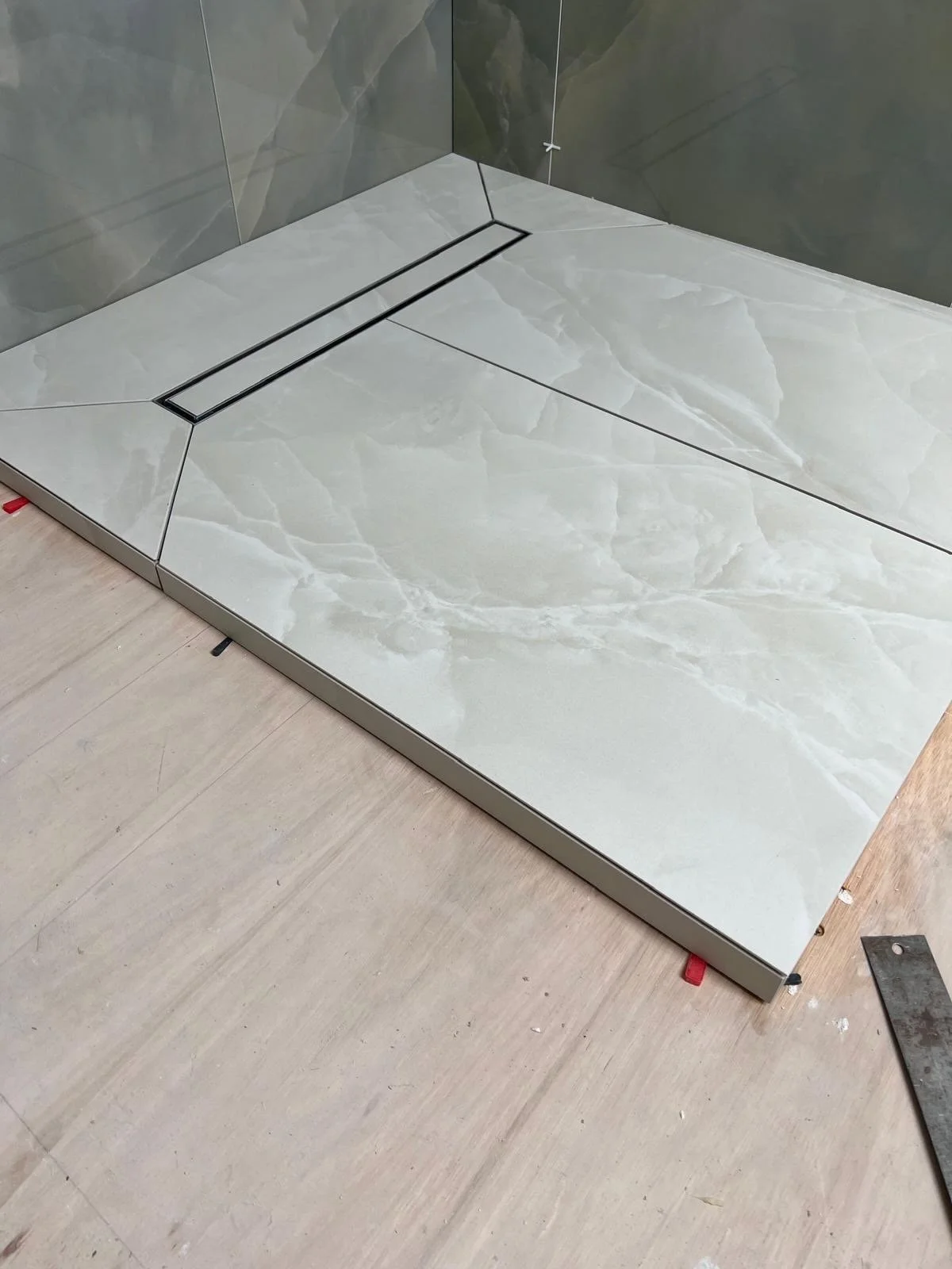 Construction site with a glass shower base installed on a wooden floor, with tiled walls in the background.