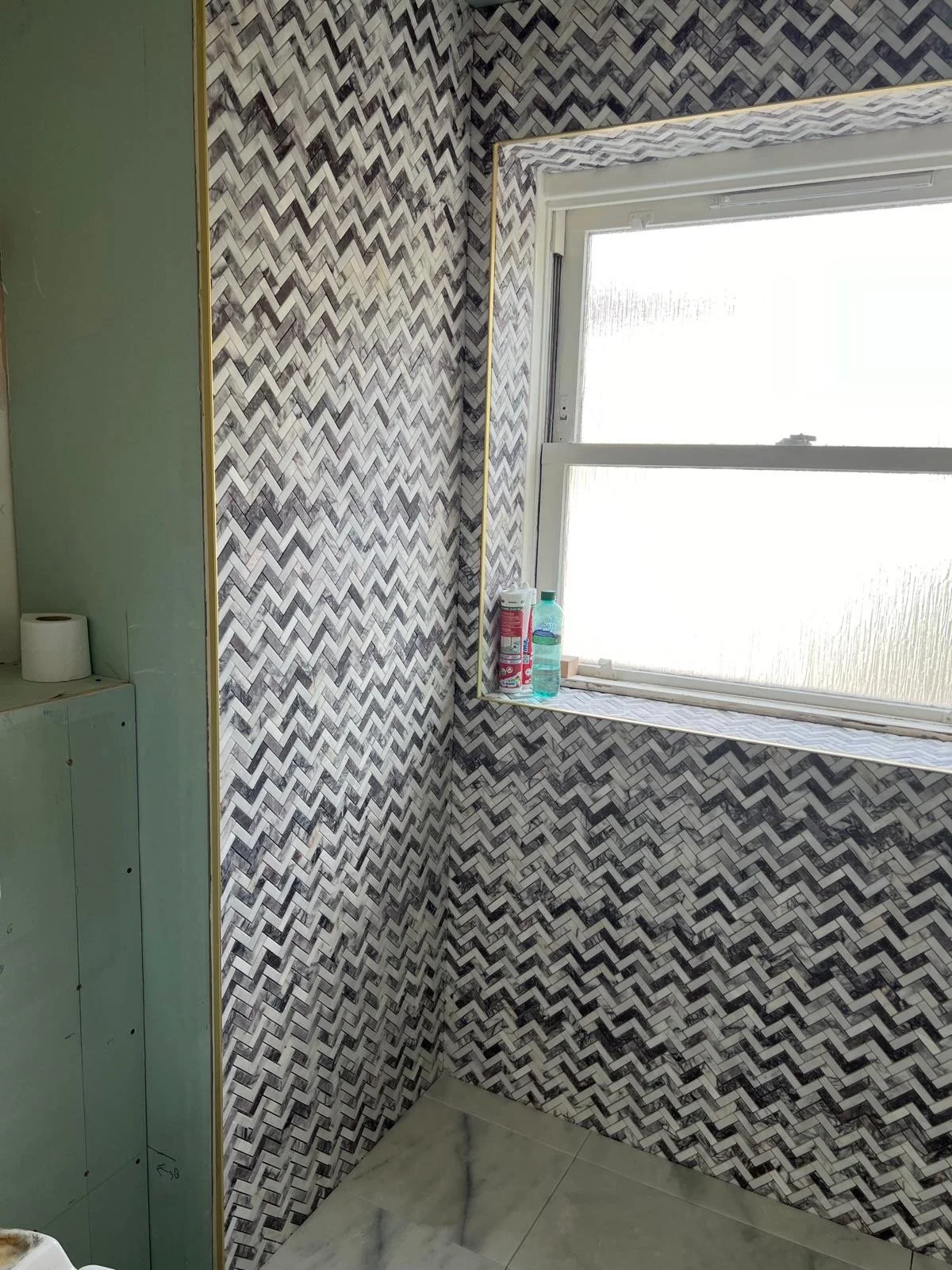 A corner of a room with patterned black, white, and gray tile walls, a window with frosted glass, and a green wall with a small section of drywall visible. On the window sill, there are some cleaning supplies and water bottles.