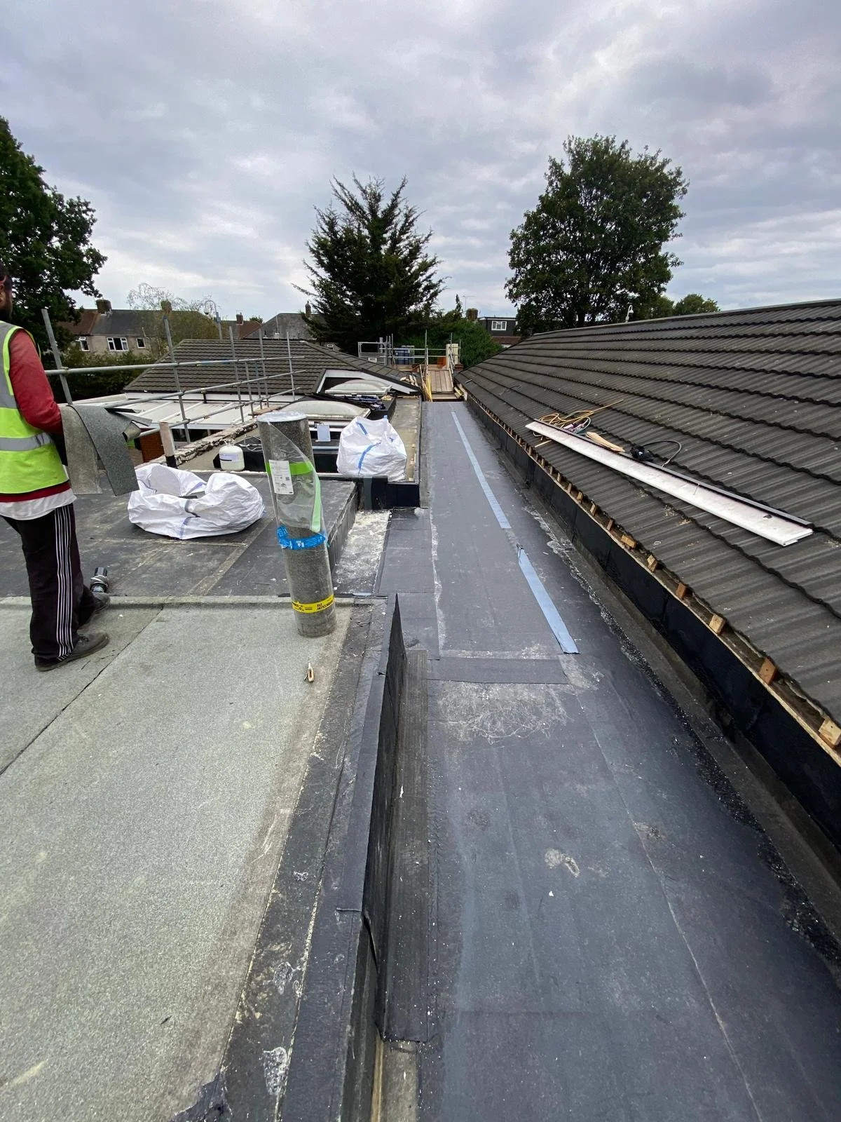 Rooftop under construction with roofing materials, tools, and a worker wearing a safety vest.