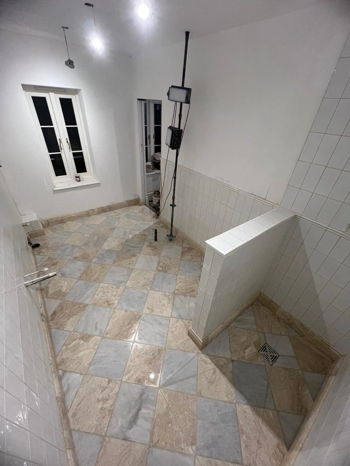 Empty room with tiled floor, white walls, and a window, with a lighting setup and some construction materials.