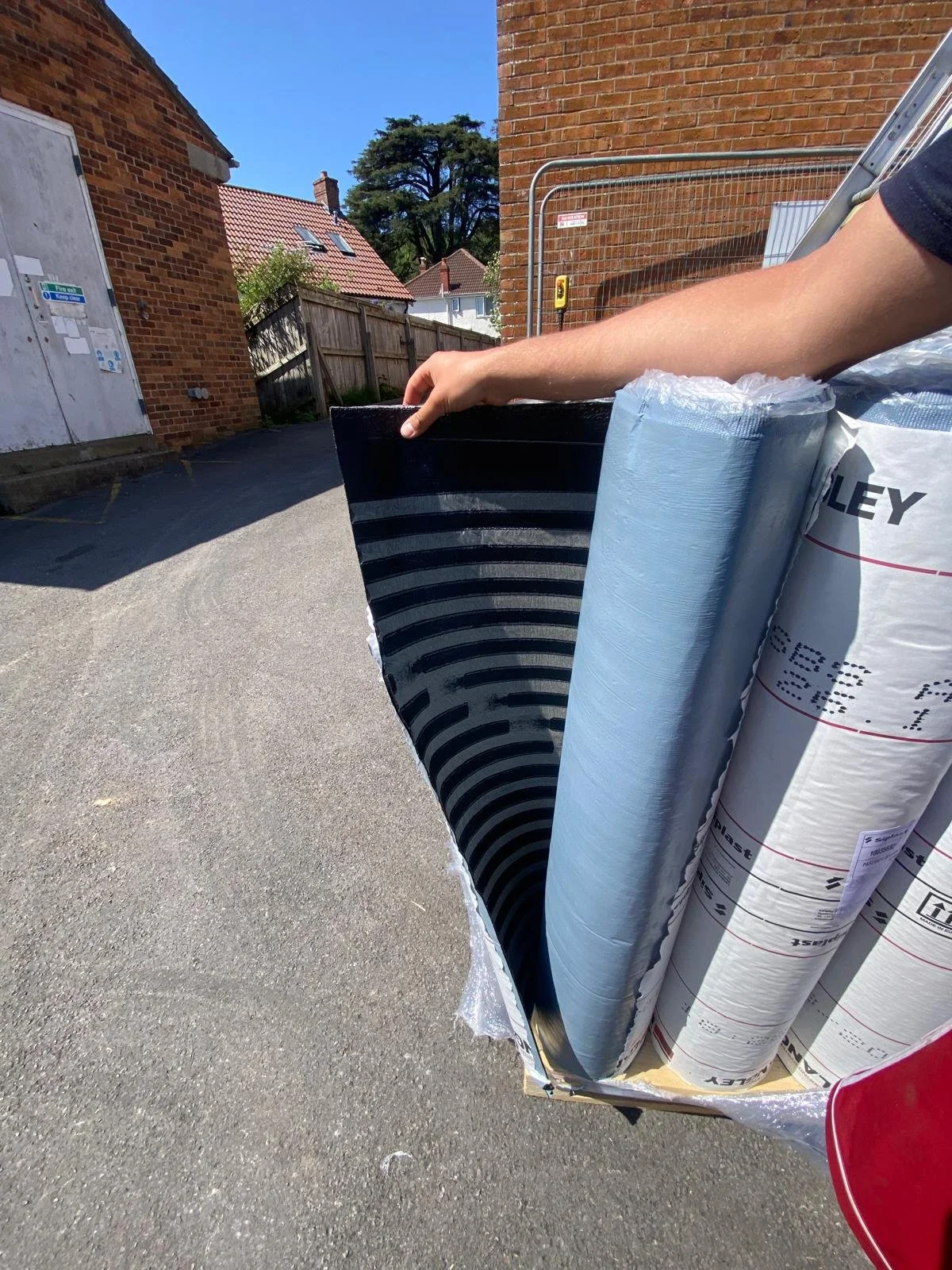 Person holding a large package of insulation rolls standing on an asphalt driveway outside near a brick house and fence.