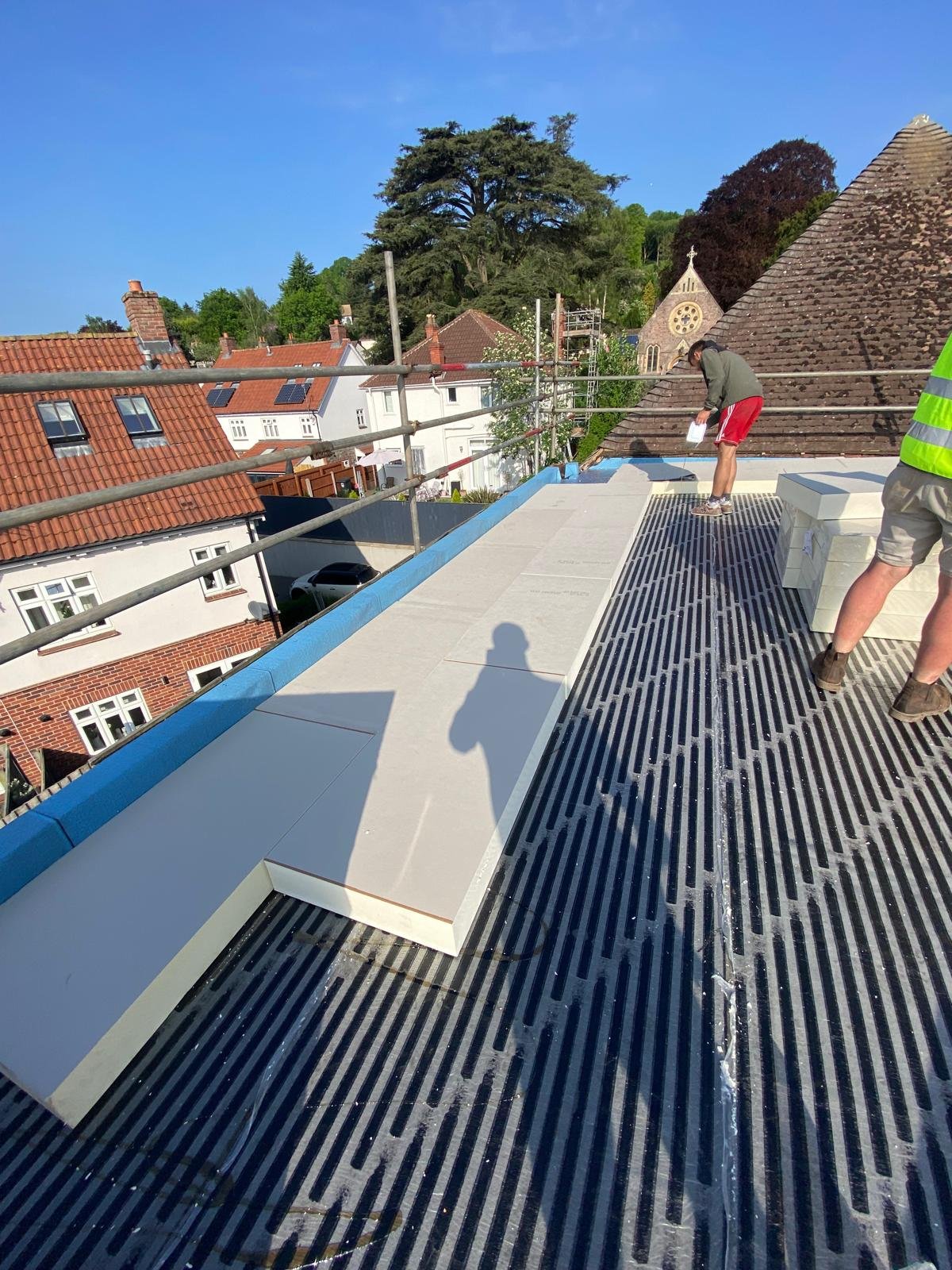 Workers installing white roofing panels on a building's rooftop during daytime, with surrounding houses, trees, and a church steeple in the background.