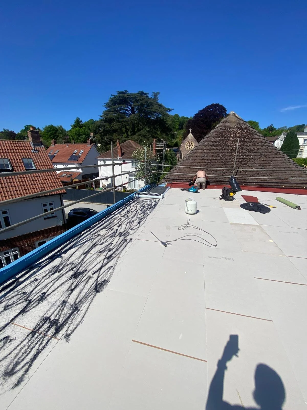 A construction worker installing roofing on a house, with tools and materials on the roof and nearby.