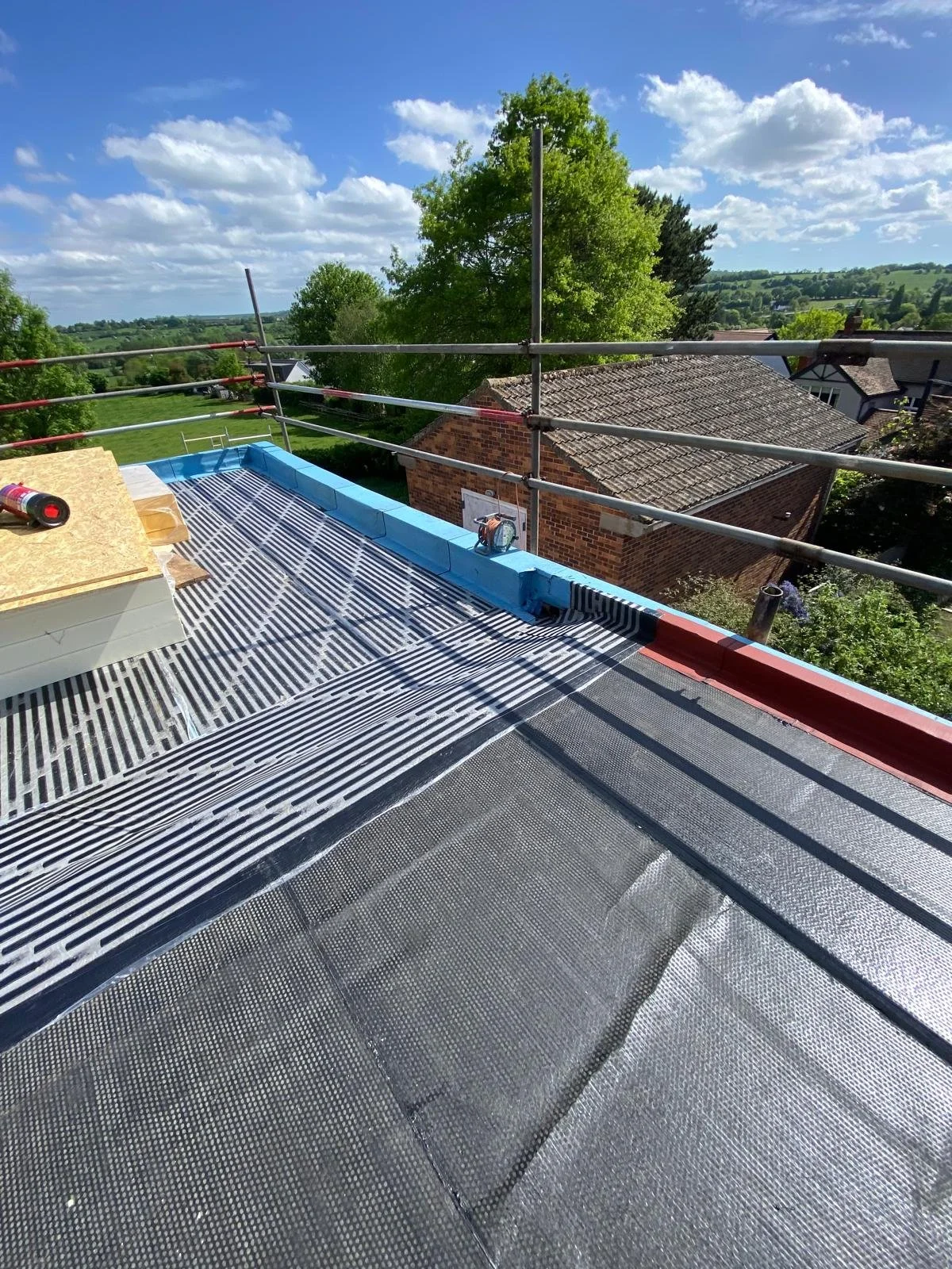 View from a rooftop under construction, with scaffolding and roofing materials, overlooking a neighborhood with trees and houses on a sunny day.