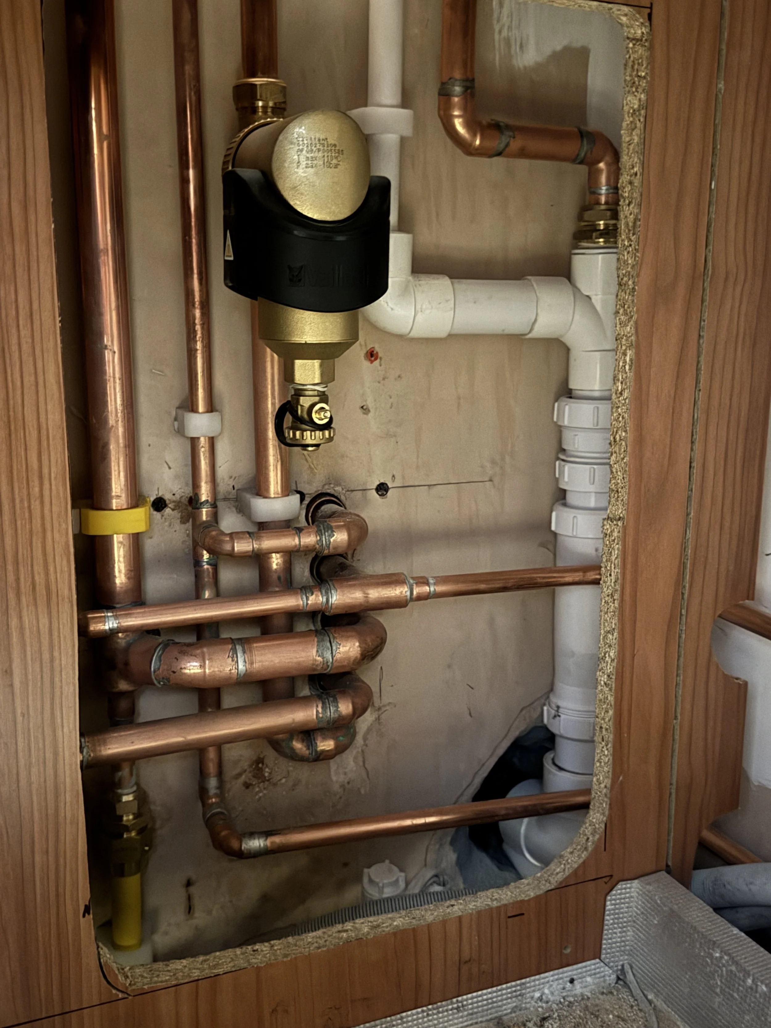 Close-up view of copper plumbing pipes and PVC pipes installed in a wooden kitchen cabinet, with some pipes wrapped in insulation.