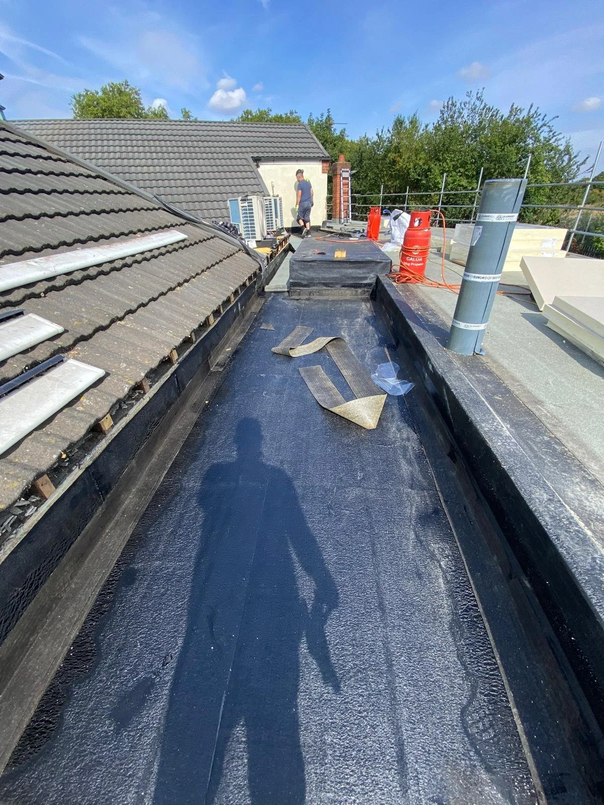 Roof work under construction, showing a flat black waterproof membrane, building tools, and two workers in the background.