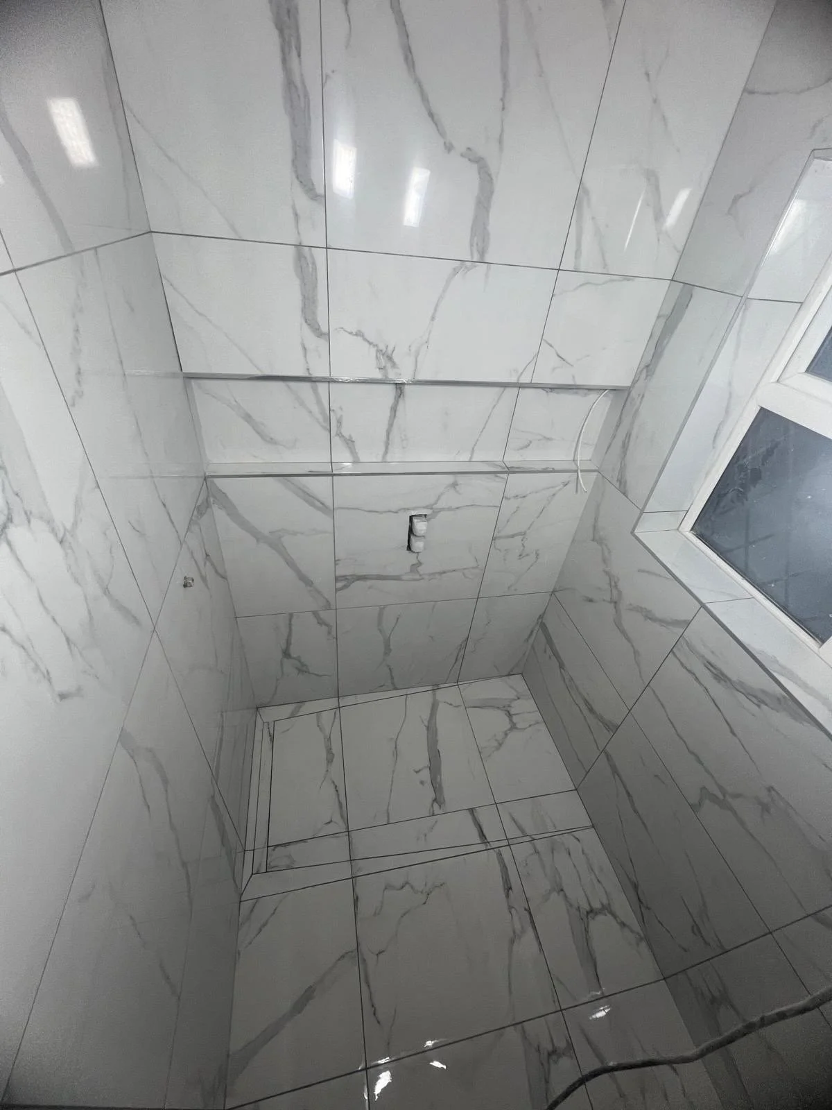 Empty room with marble-like tiled walls and floor, a small hole in the wall, and a window on the right side.