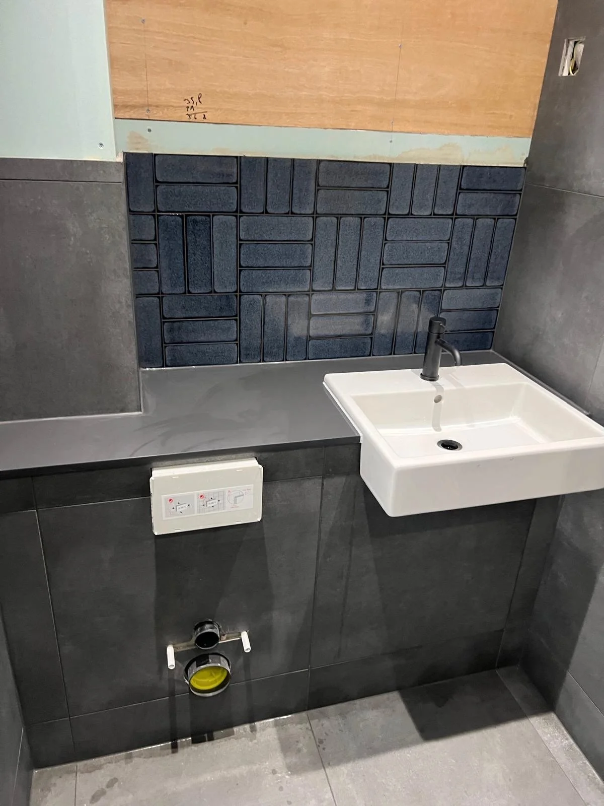 Indoor bathroom with gray tiled walls, black and white sink, and a metal countertop. The wall behind the sink has a section with dark blue tiles, and there is an electrical outlet and a plumbing pipe beneath the sink.