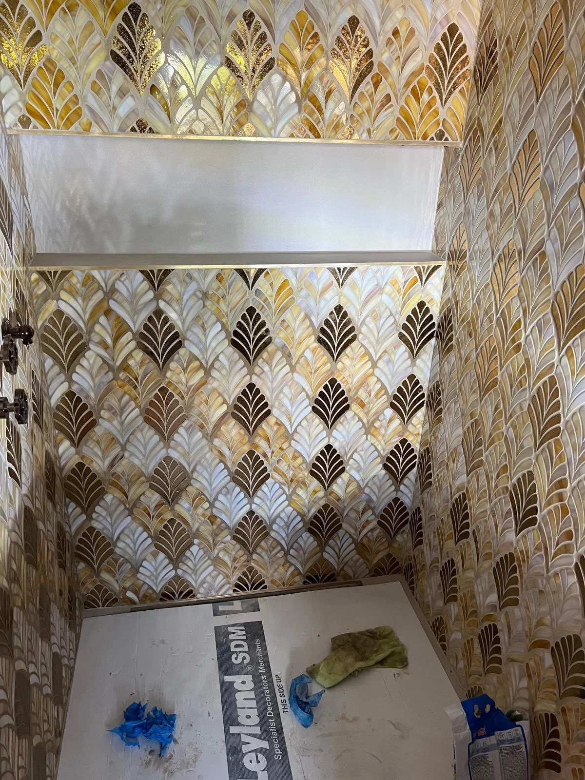A bathroom shower area with a gold, white, and black art deco tile pattern on the walls, a white shelf at the top, and a floor with some cleaning cloths and tools.