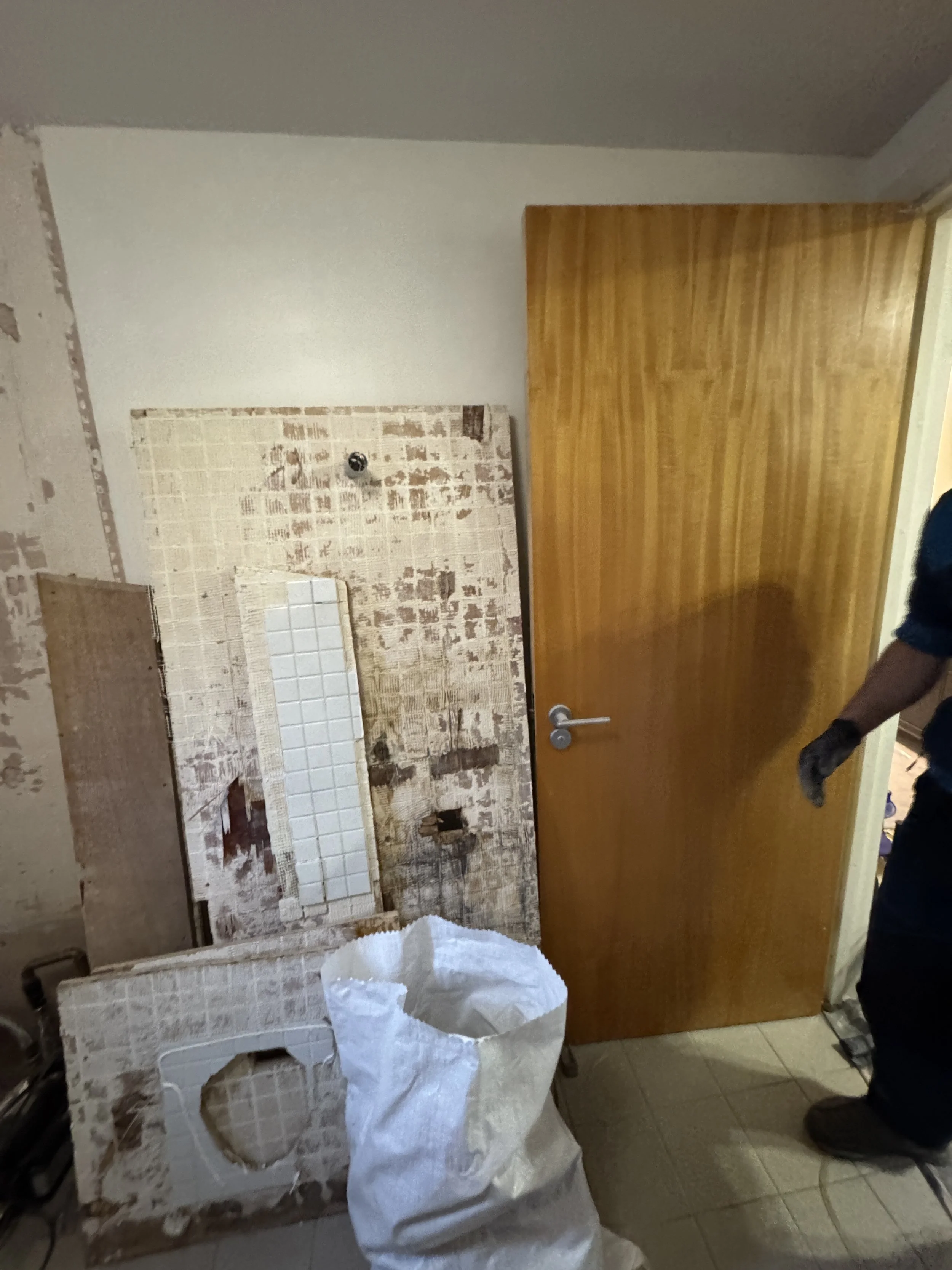 A room with a partially removed tiled wall, a wooden door, and a waste bag on the floor.