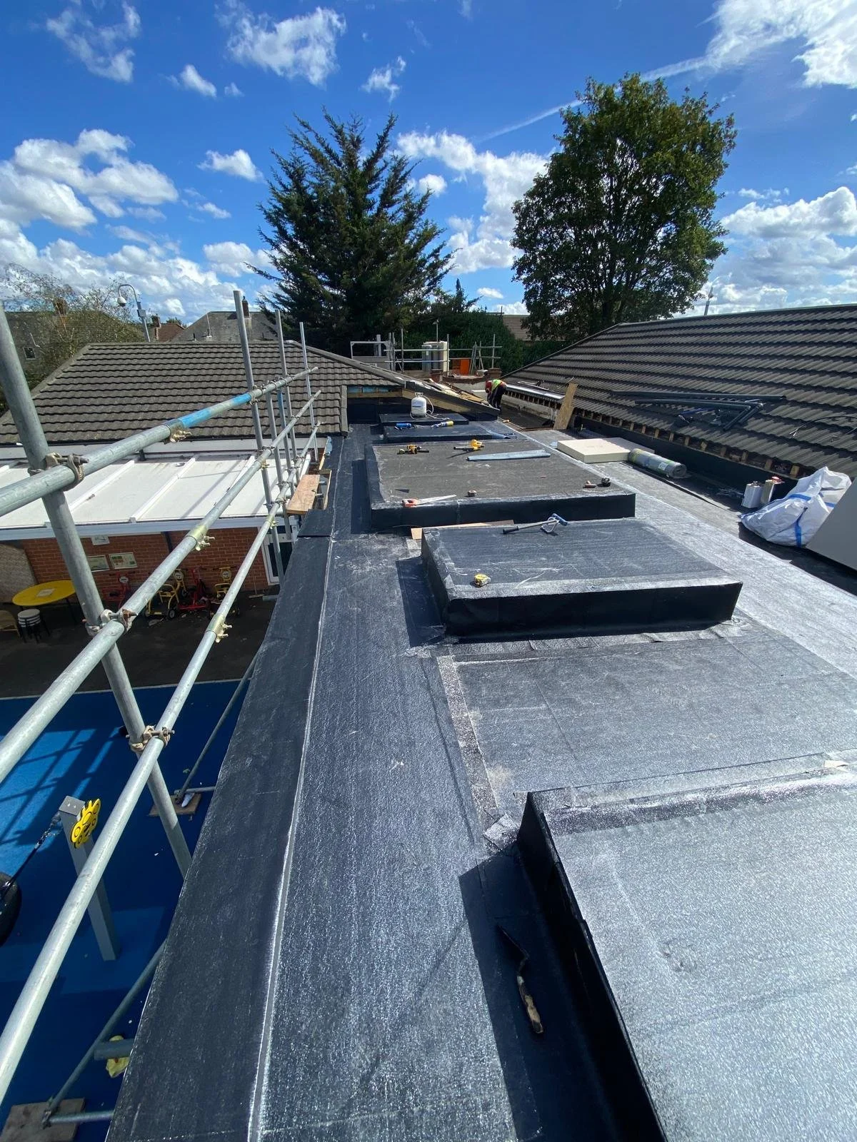 Construction workers installing roofing on a building, with scaffolding around the roof and a blue sky with scattered clouds overhead.