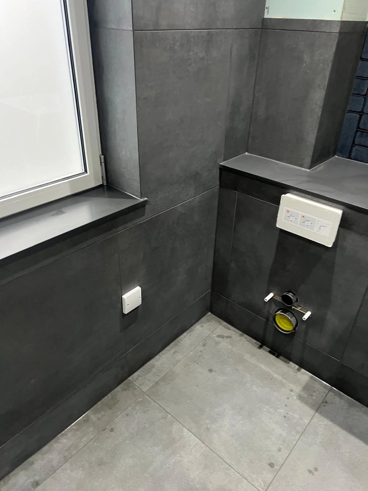 The corner of a modern bathroom with dark gray tiled walls and light gray tiled floor. There is a window sill, a switch on the wall, and plumbing fixtures on the wall for a toilet.