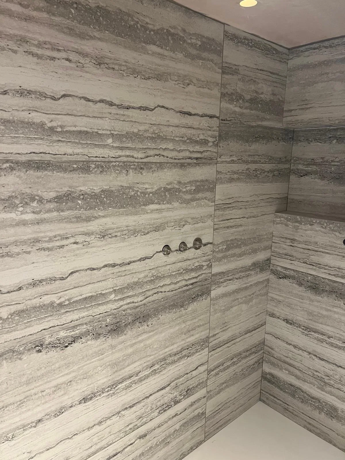 Interior view of a bathroom shower with large, horizontally striped gray and beige tiles and three showerhead fixtures on the wall.