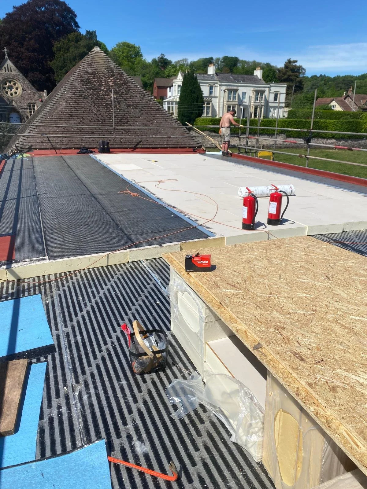 A construction worker on a roof under construction with safety railings, fire extinguishers, and building materials, with neighboring houses and trees in the background.